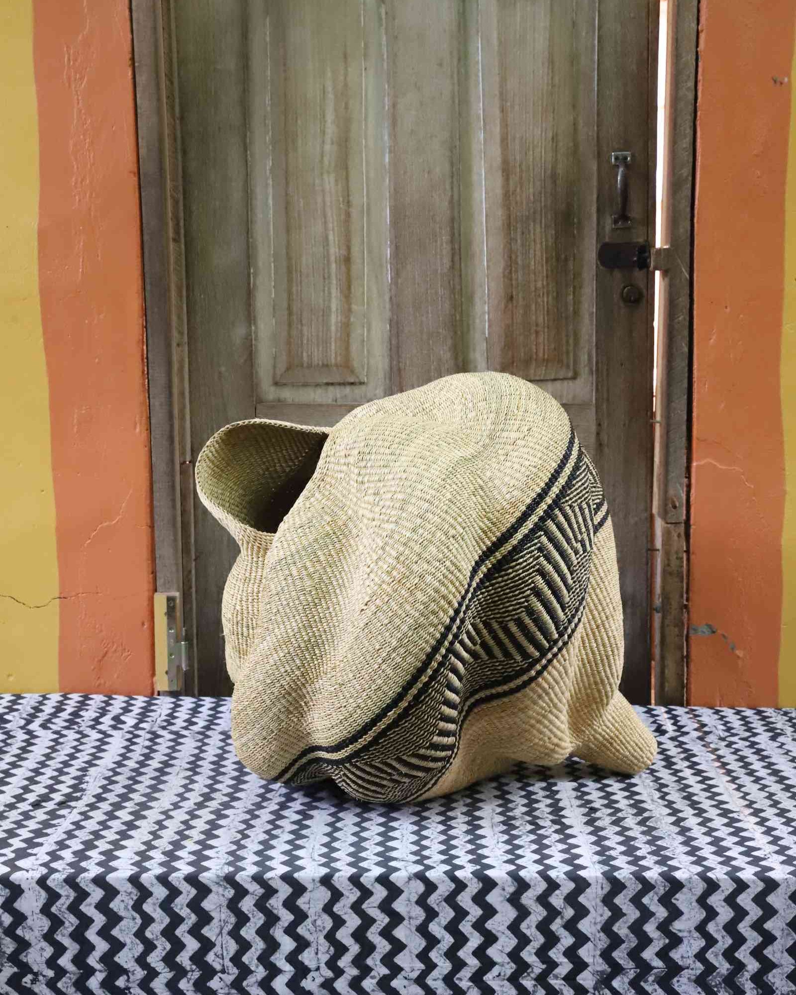 Sculptural spiral-form Bolga basket in natural straw with a black geometric band, styled on a patterned tablecloth.