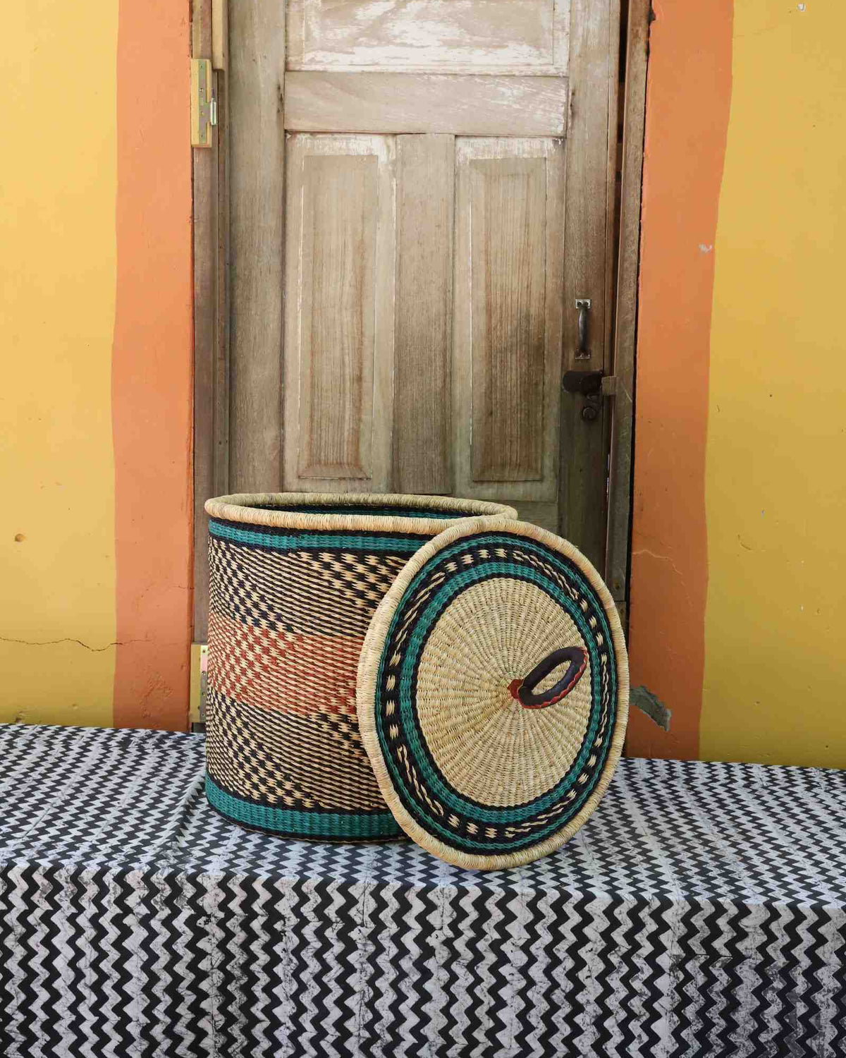 Handwoven Bolga laundry basket with lid removed, natural body with orange bands and teal rim accent.