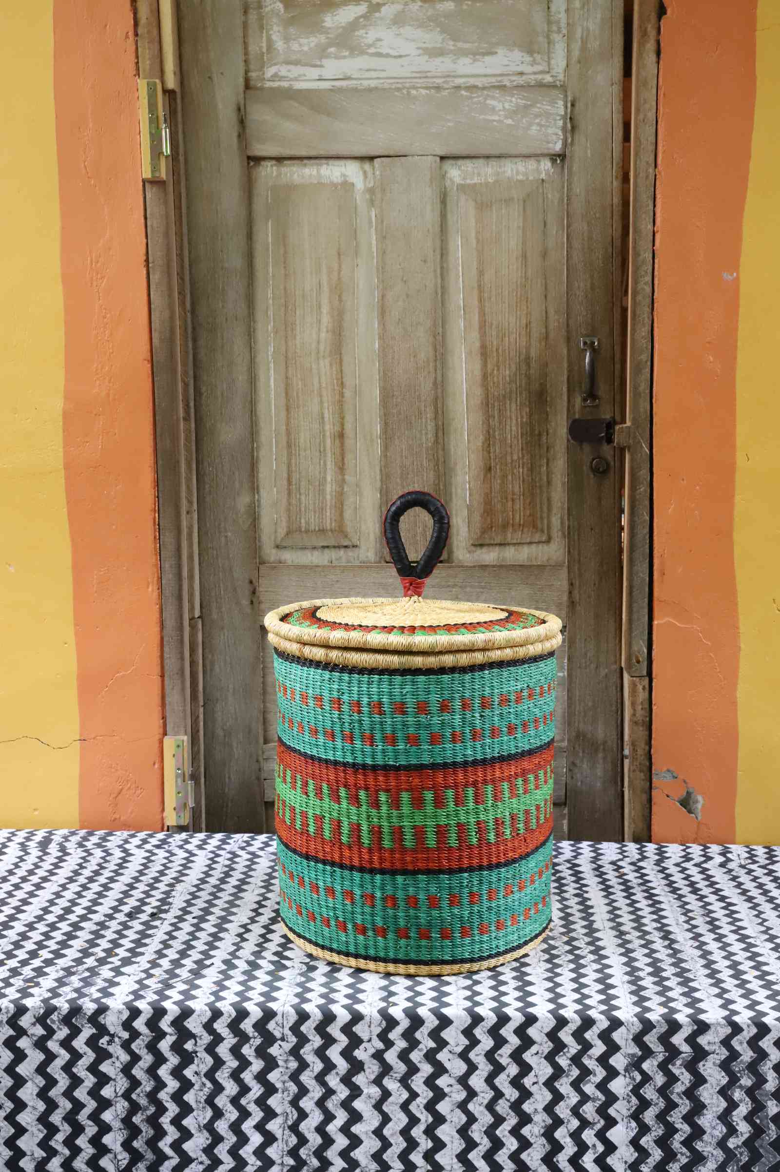 Handwoven Bolga laundry basket with coiled rim and lid in natural straw with teal, red and green banded stripes.