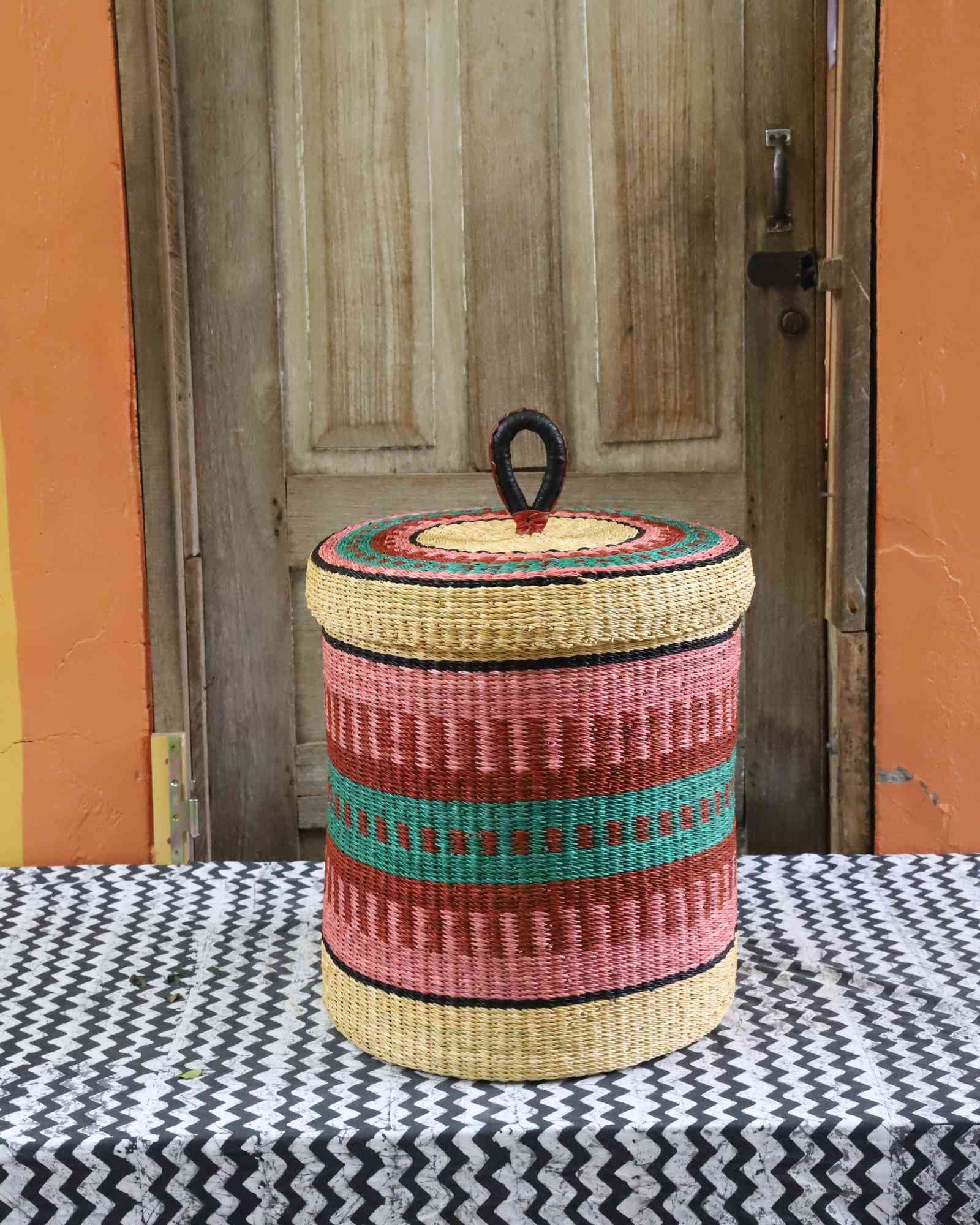 Lidded laundry basket in natural straw with pink and teal striped banding, styled on a chevron cloth by a wooden door.