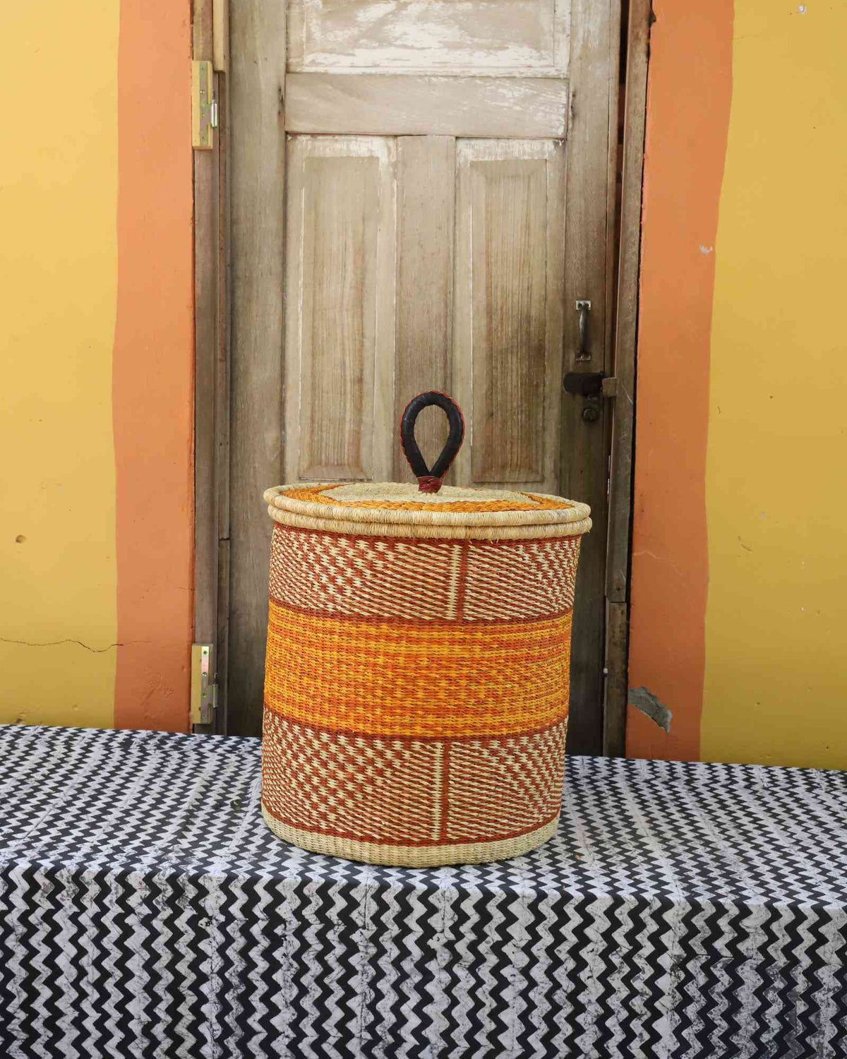 Handwoven Bolga laundry basket with coiled rim and lid in natural straw with an orange central band.