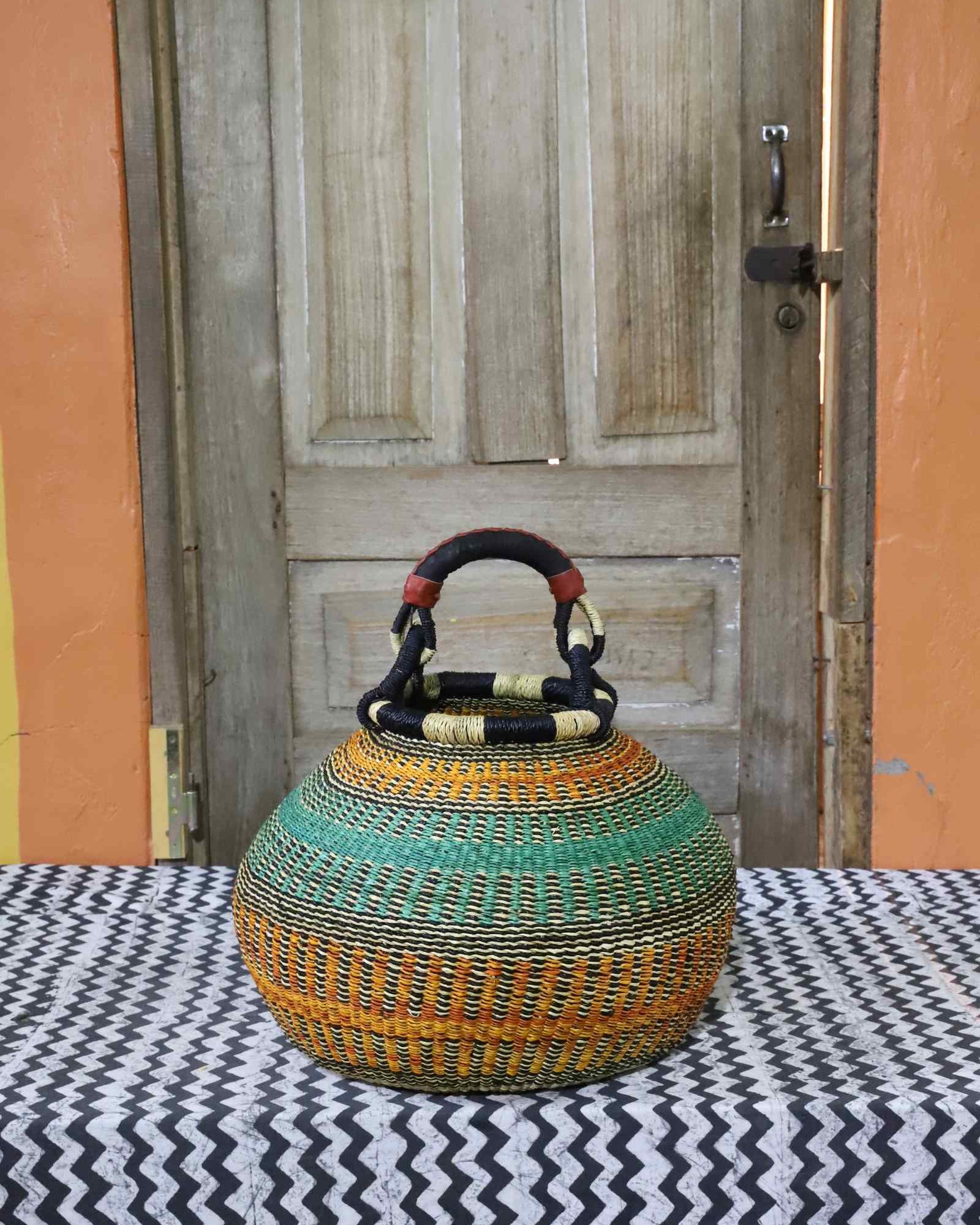 Round Bolga pot basket in orange and green stripes with a leather-wrapped arched handle, on a tablecloth.