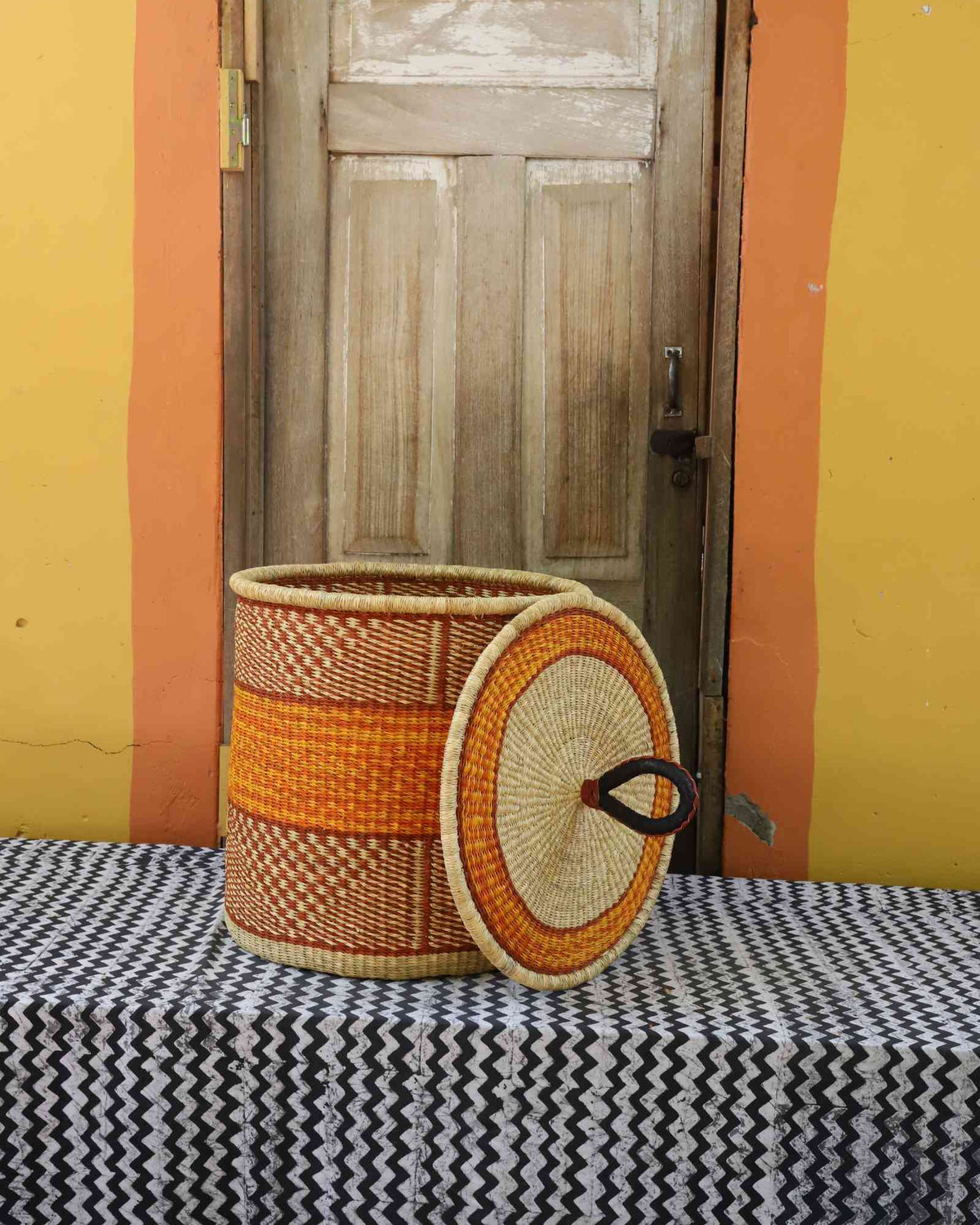 Handwoven Bolga laundry basket with lid removed, natural body with orange central band and coiled lid.