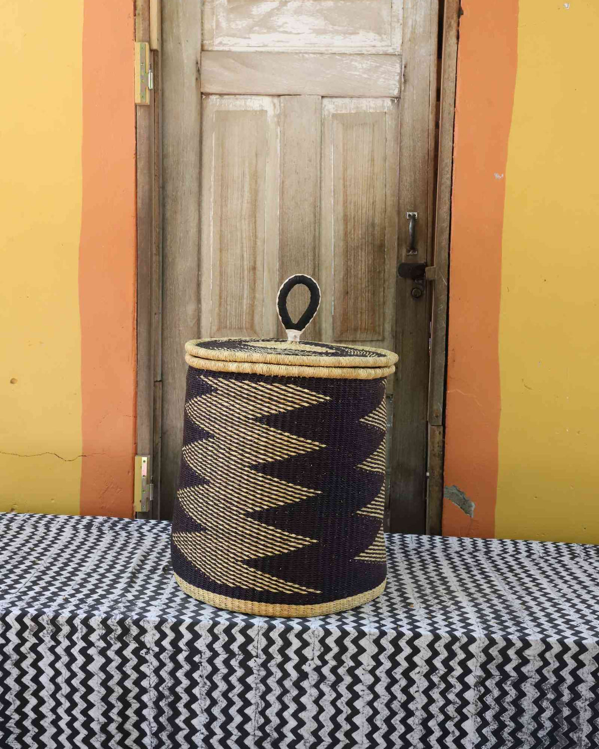 Handwoven Bolga laundry basket with coiled rim and lid in natural straw with a bold black zigzag lower band.