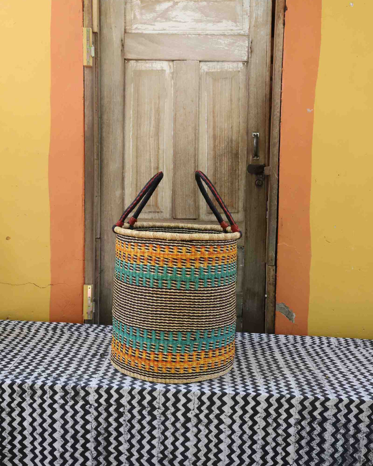 Medium handwoven open-top Bolga laundry basket in natural straw with orange, teal and green banded stripes.
