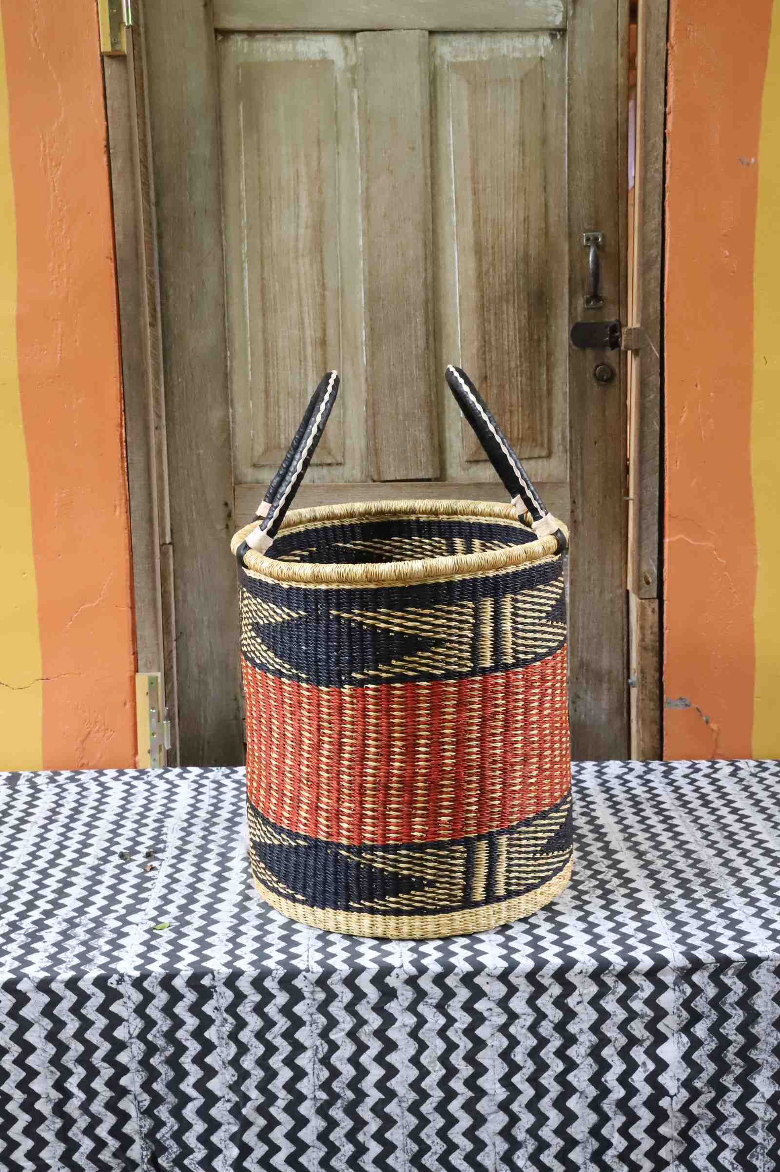 Open laundry basket in natural straw with an orange and black zigzag band, styled on a chevron cloth by a wooden door.