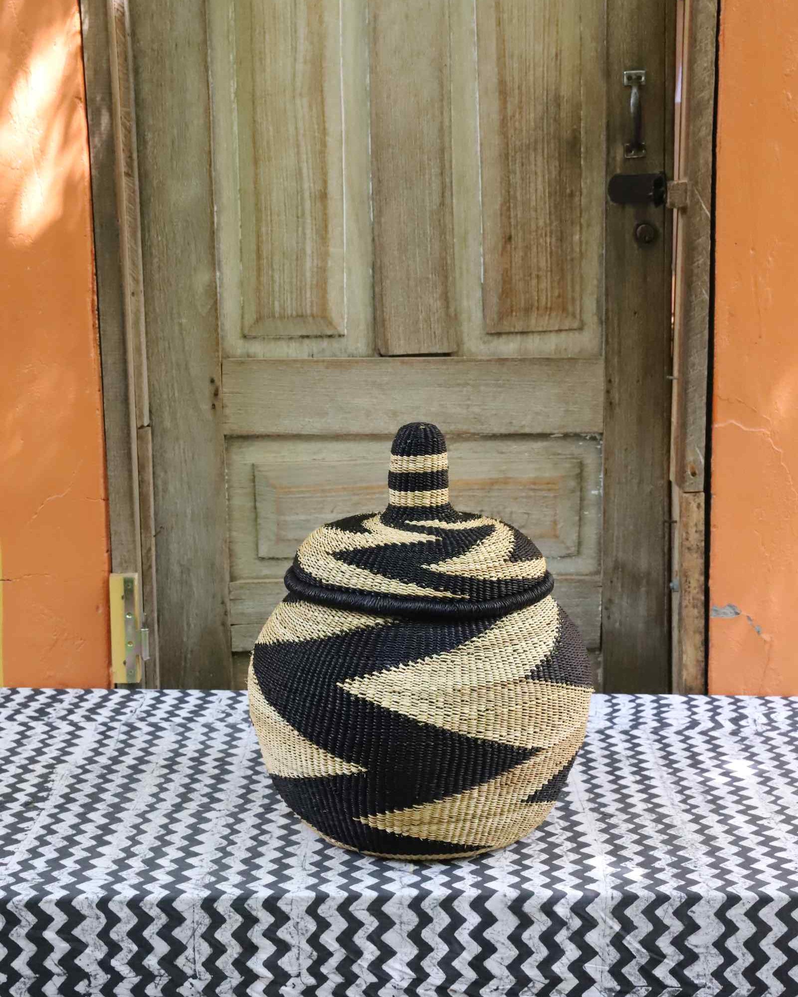 Lidded Bolga storage basket with a bold black and natural zigzag pattern and a domed fitted lid, on a tablecloth.
