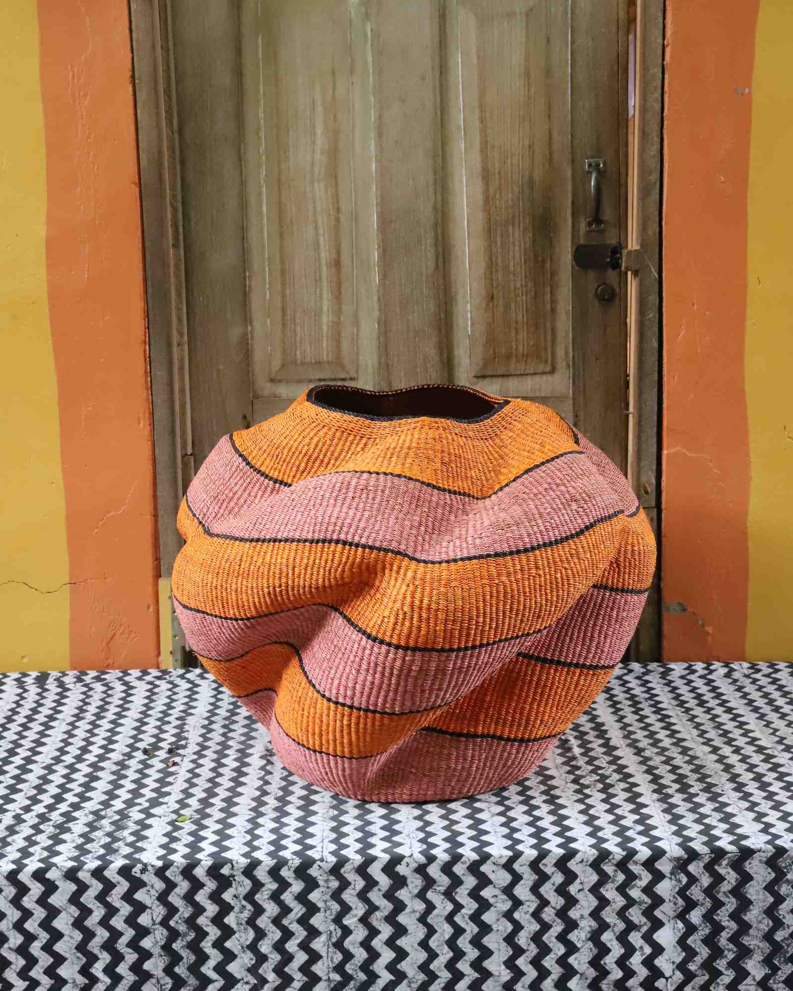 Large nest-shaped Bolga art basket in pink and rust wavy braided weave, styled on a chevron cloth by a wooden door.