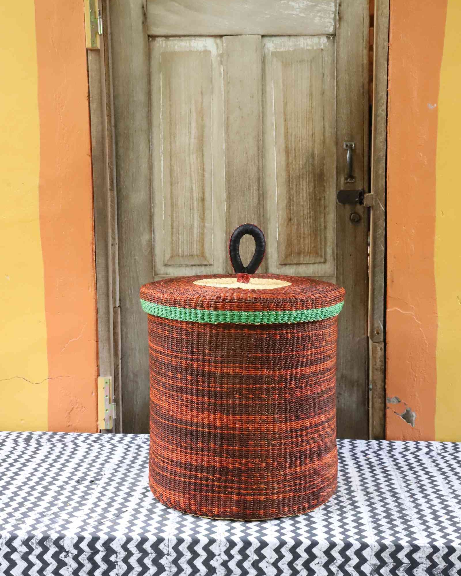 Handwoven Bolga laundry basket with lid in rust-orange elephant grass with a green banded rim.