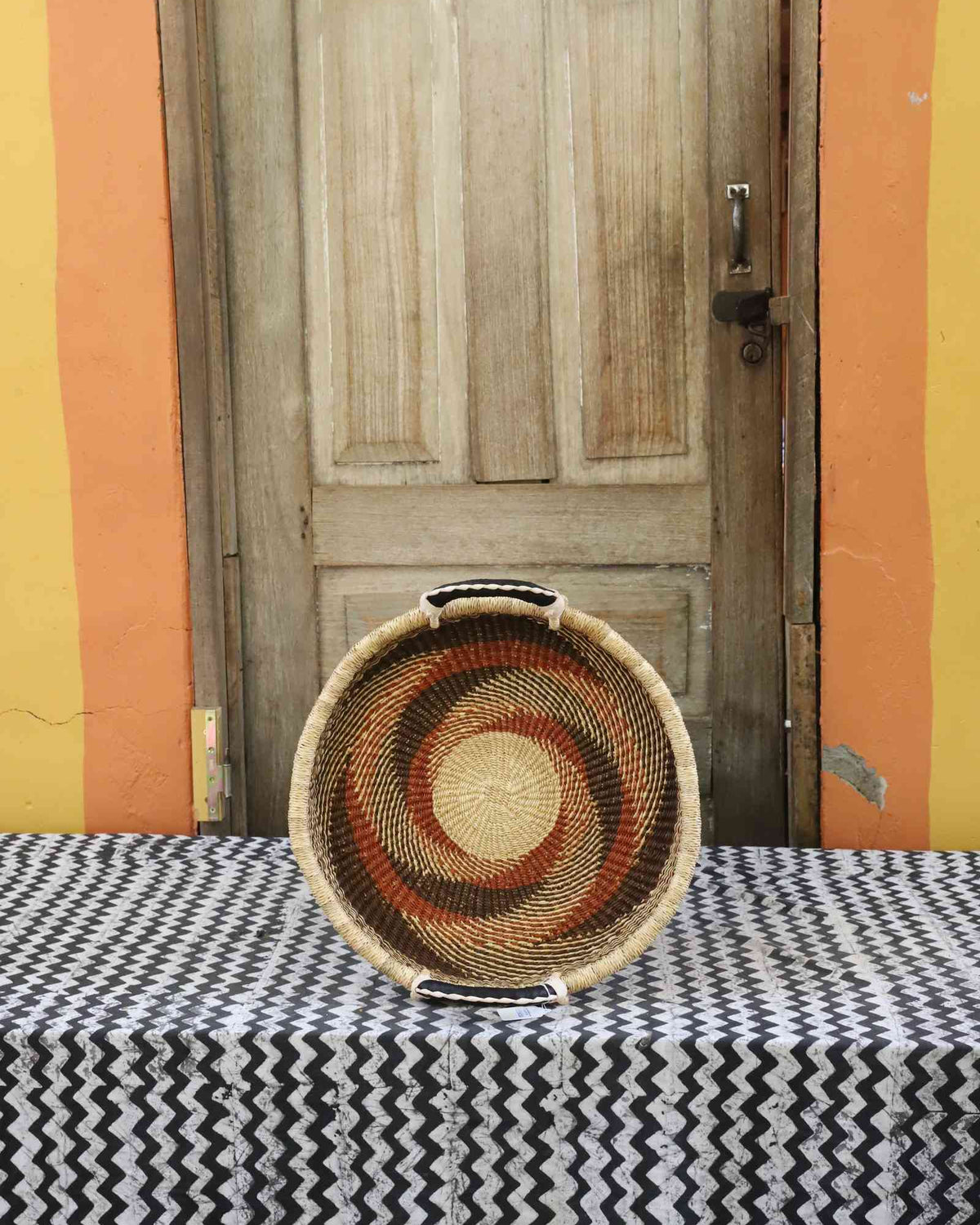 Handwoven Bolga wall tray disc in natural straw with rust-red pinwheel swirl and yellow centre.
