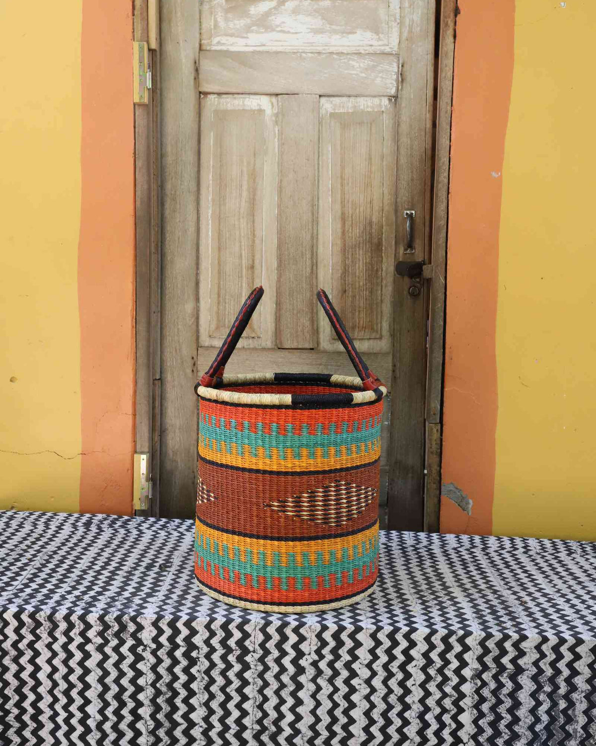 Small handwoven open-top Bolga laundry basket in dyed orange with green and red banded stripes.