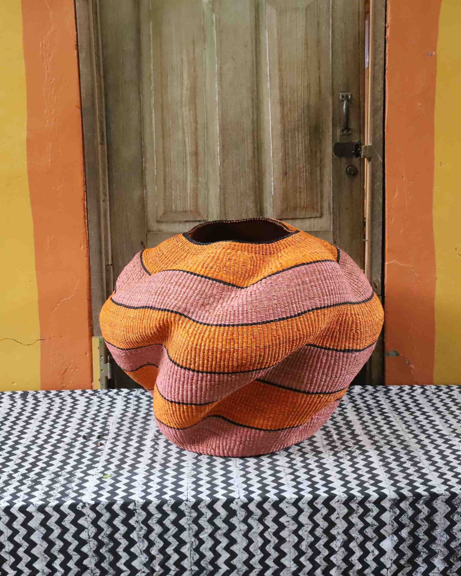 Large nest-shaped Bolga art basket in pink and rust wavy braided weave, front view on a chevron cloth.