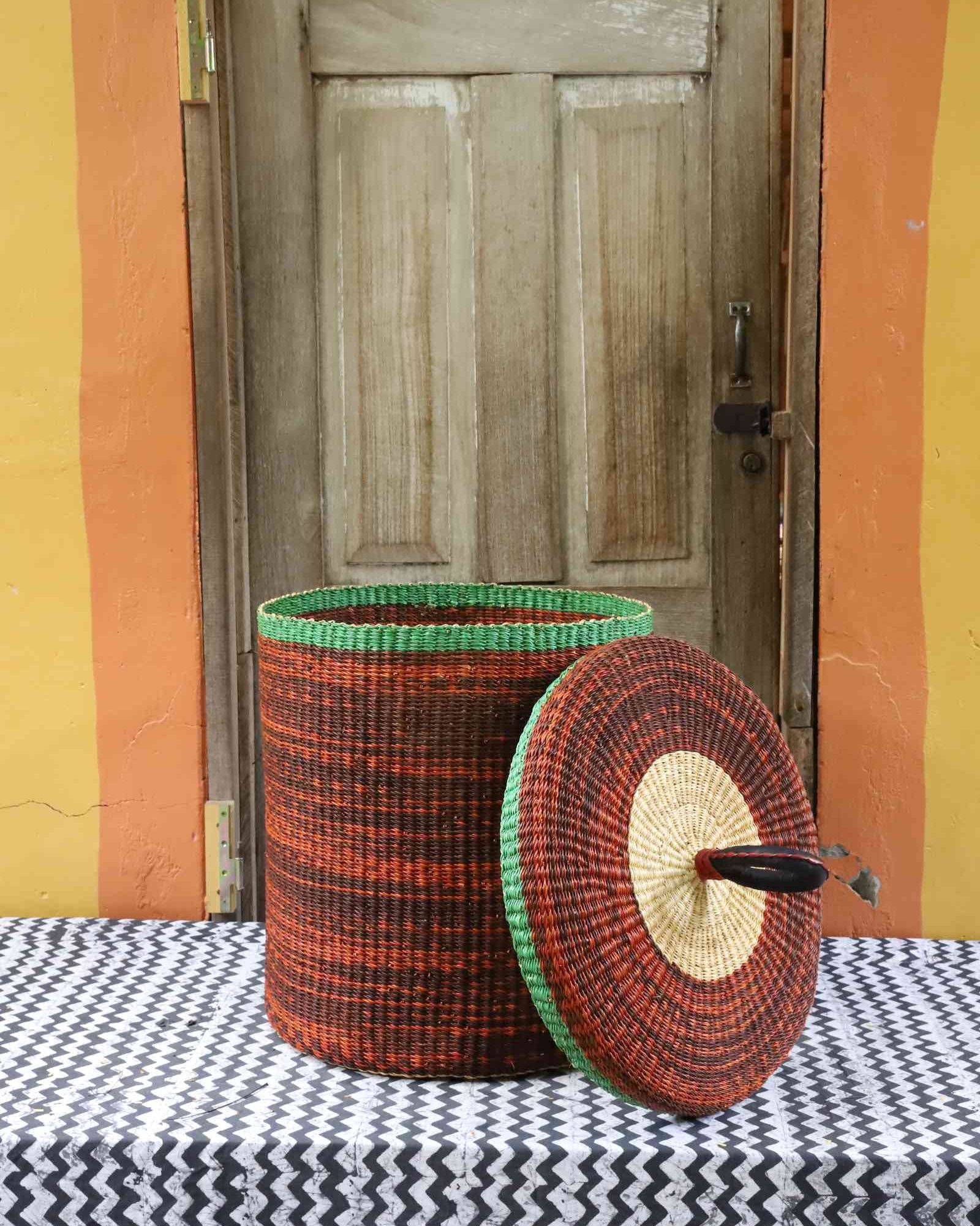 Handwoven Bolga laundry basket with lid removed, showing rust-orange body with green rim and matching lid.