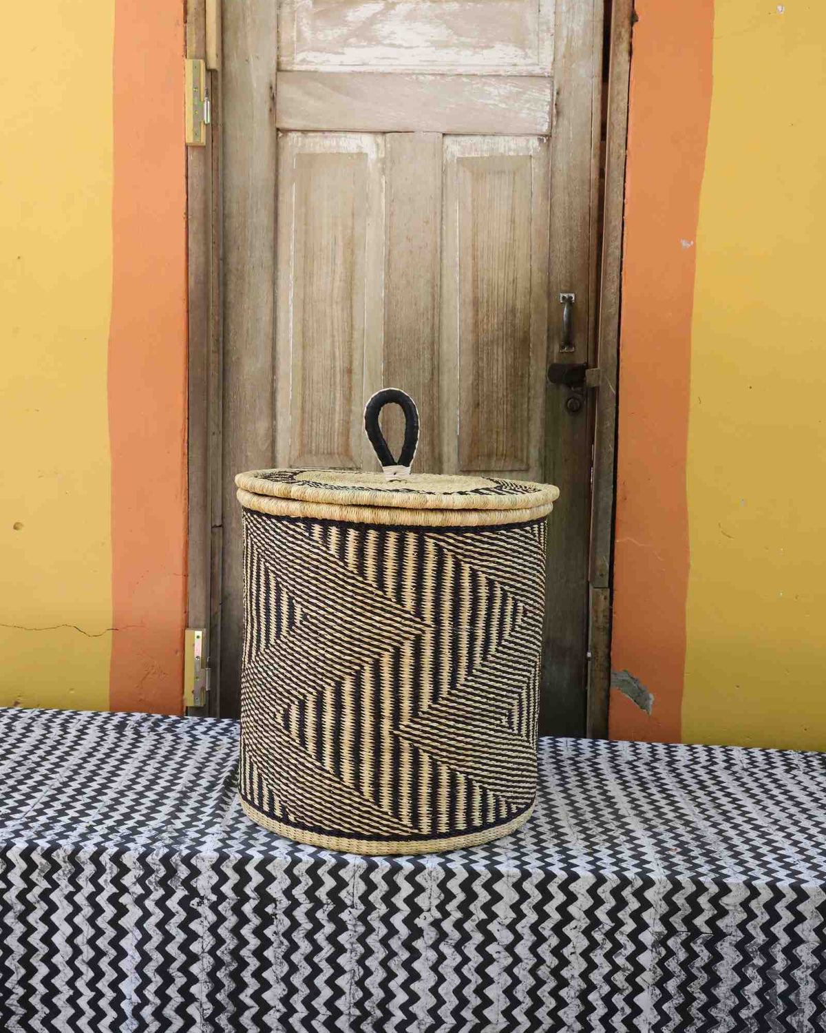 Handwoven Bolga laundry basket with coiled rim and lid in natural straw with dark diagonal wave stripes.