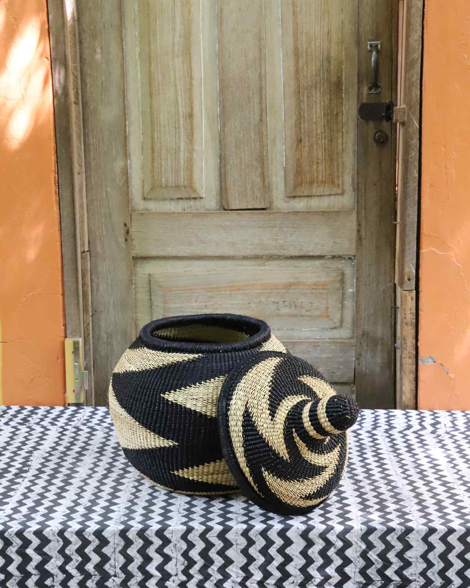 Lidded Bolga storage basket in a black and natural zigzag pattern, shown with its domed lid removed beside it.