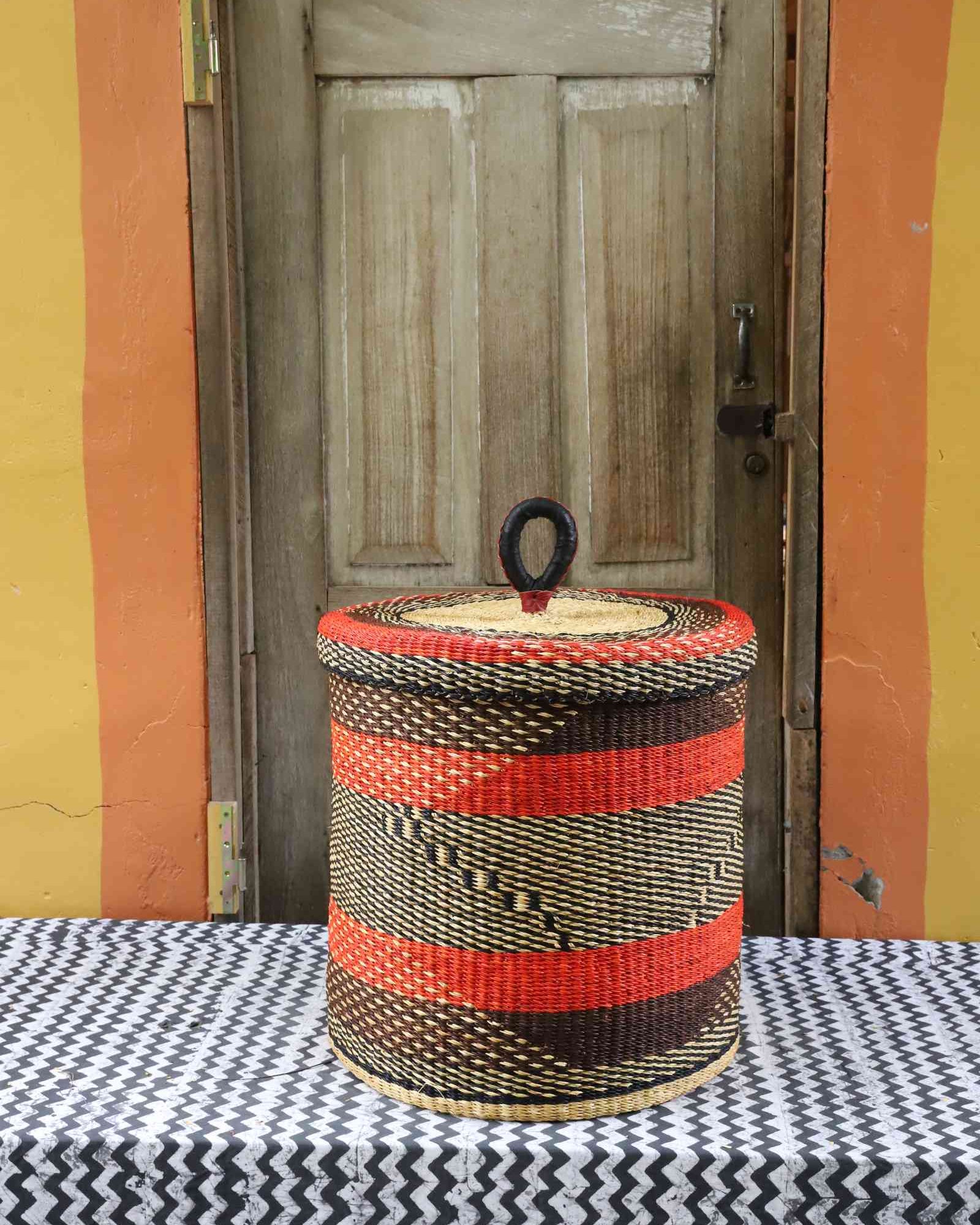 Handwoven Bolga laundry basket with lid in natural straw with red-orange stripes and a black diamond weave band.