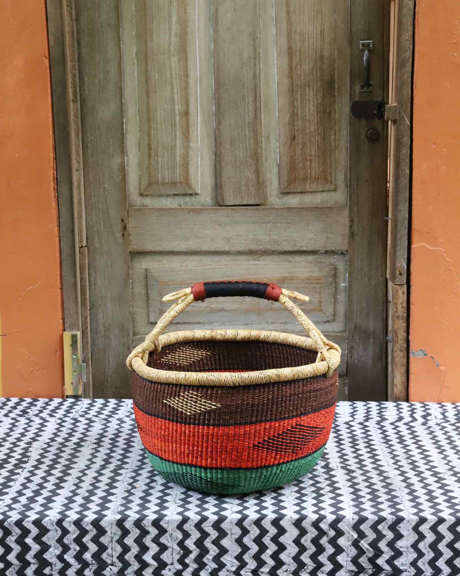 Round Bolga shopping basket with a leather-wrapped handle, striped in brown, orange and green, on a tablecloth.