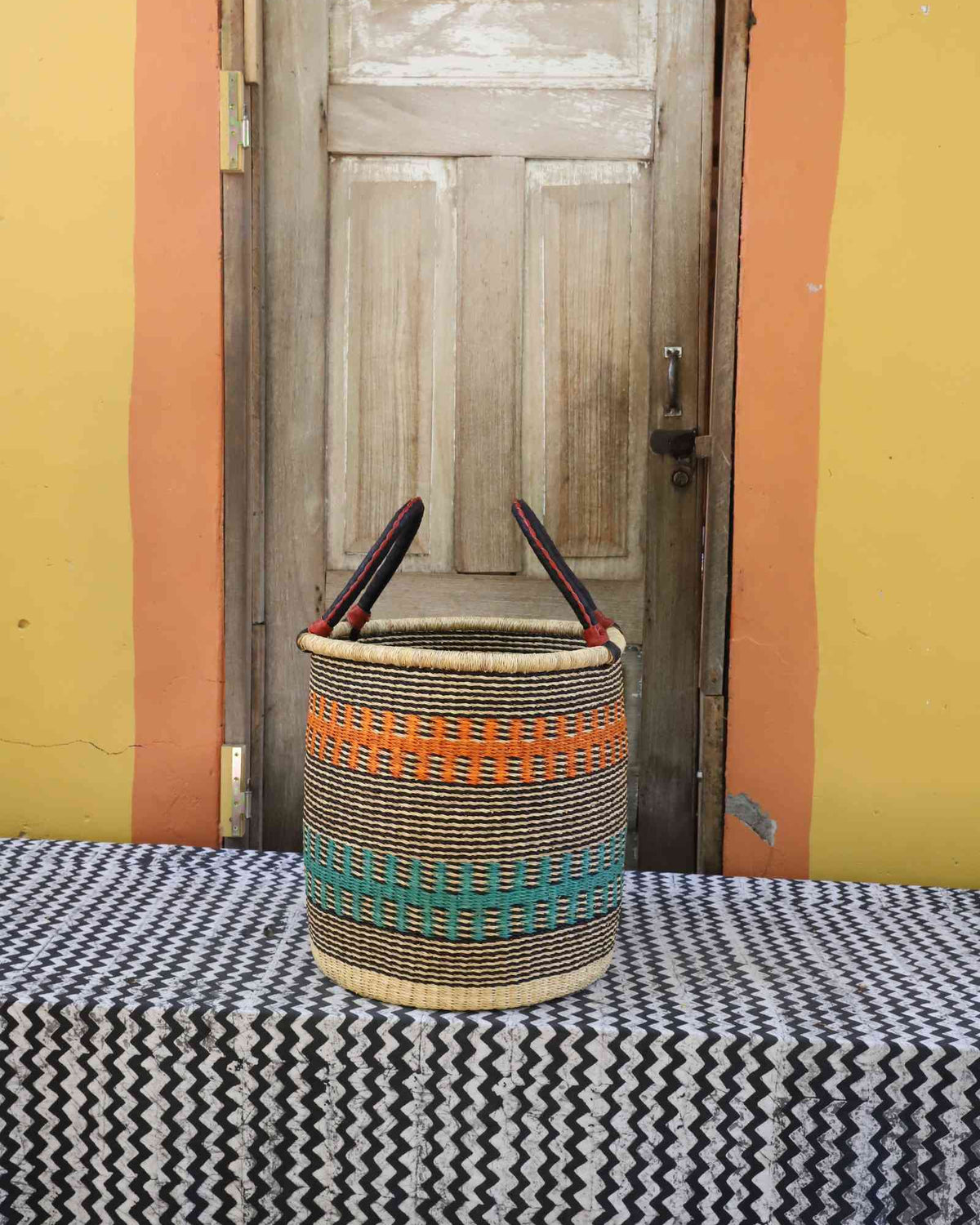 Small handwoven open-top Bolga laundry basket in natural straw with orange and teal banded stripes.