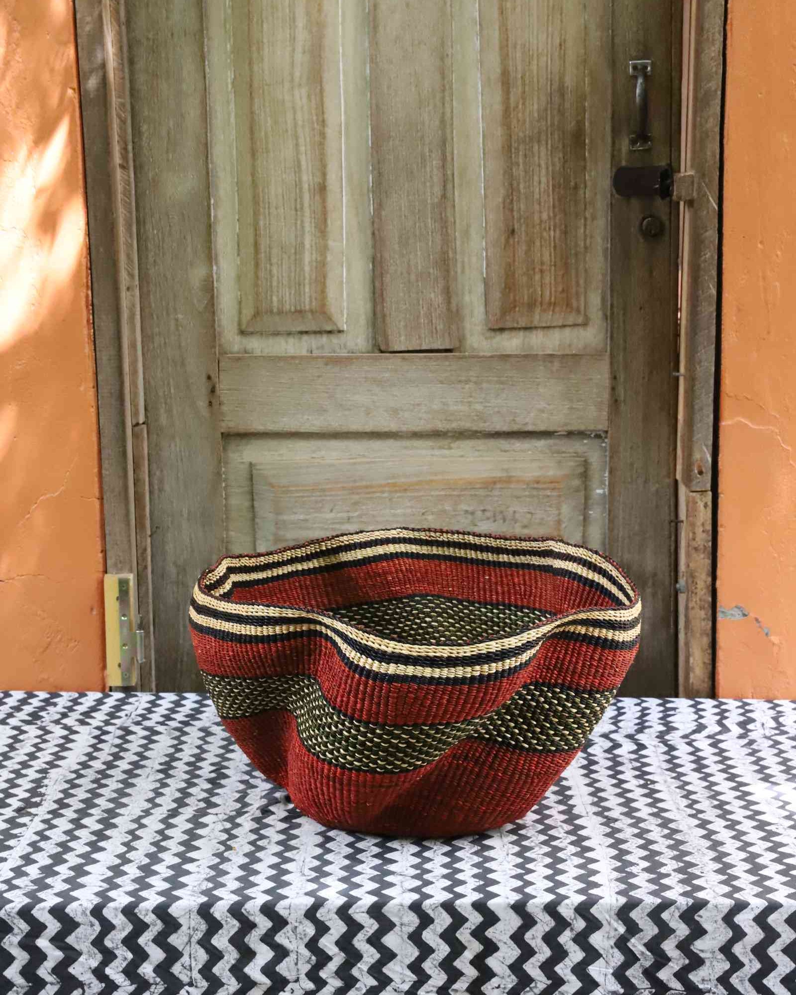Wide wave-rimmed Bolga basket in rust red with a black and gold wave-patterned band, styled on a tablecloth.