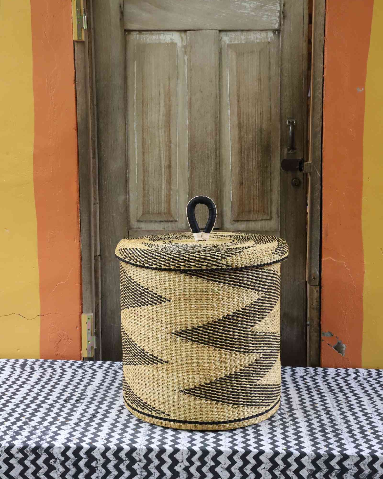 Handwoven Bolga laundry basket with lid in natural straw with a black zigzag pattern and looped lid handle.