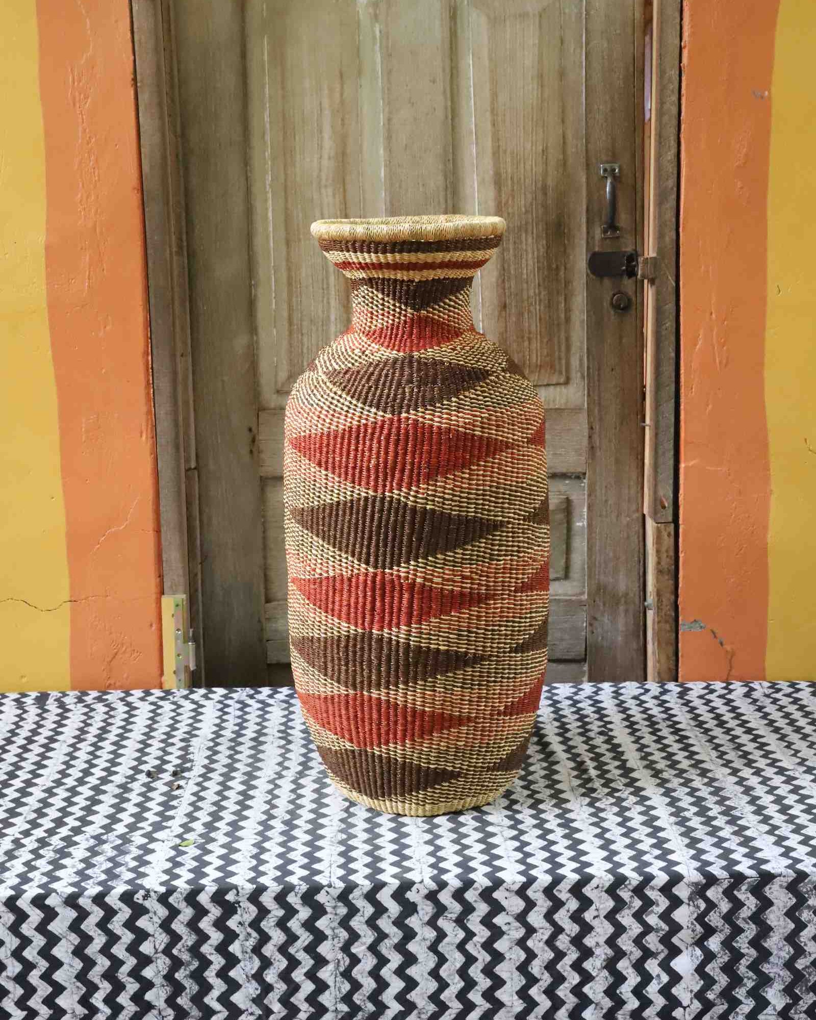 Tall vase-shaped Bolga art basket in natural straw with a brown and rust diamond pattern, styled on a chevron cloth by a wooden door.