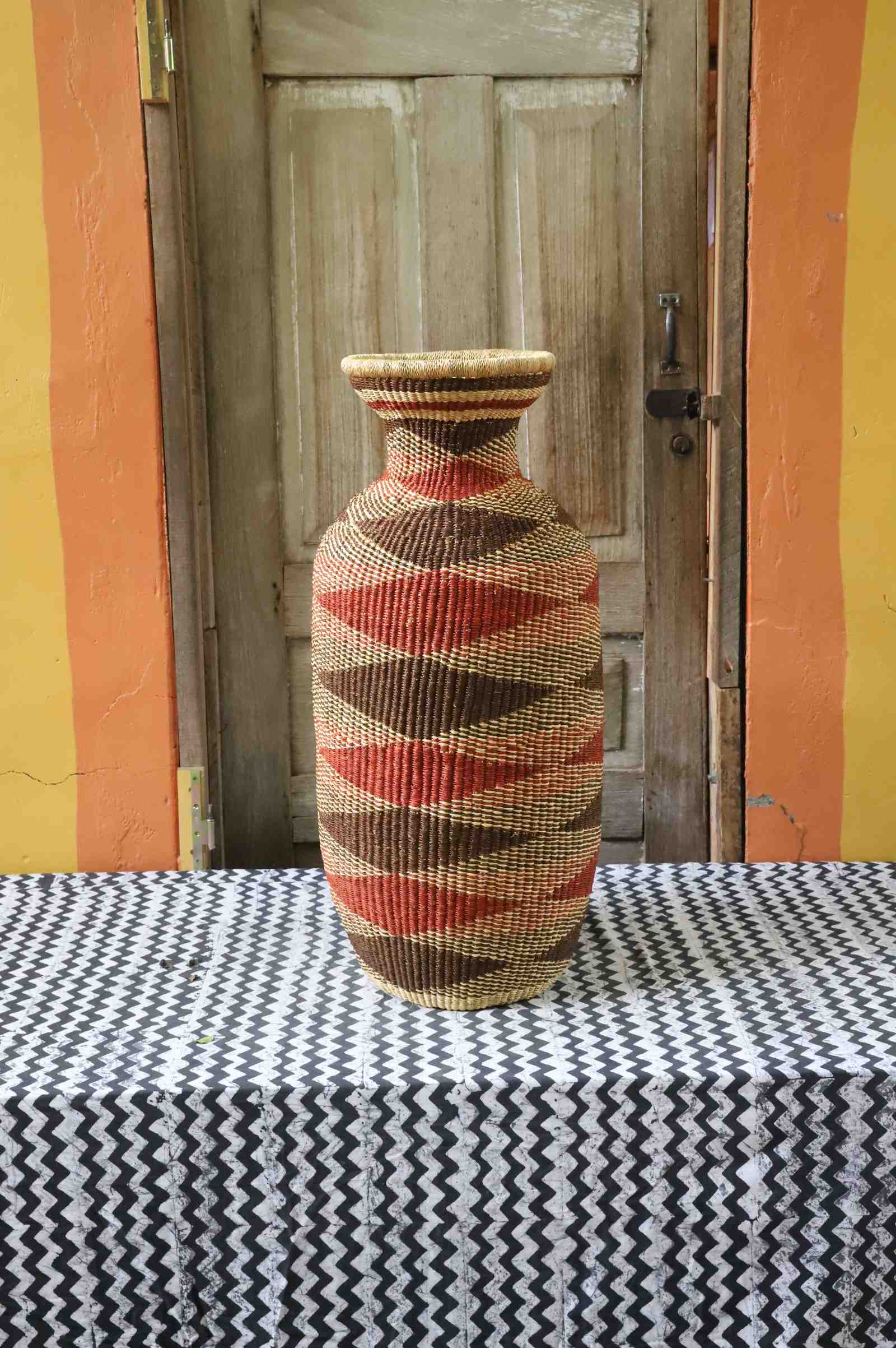 Tall vase-shaped Bolga art basket in natural straw with a brown and rust diamond pattern, styled on a chevron cloth by a wooden door.