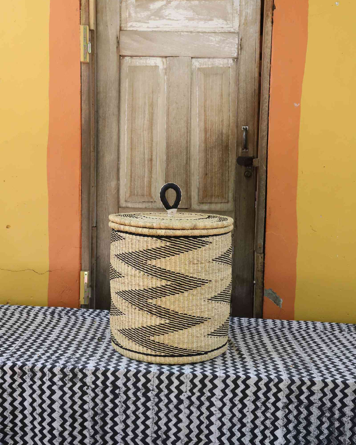 Handwoven Bolga laundry basket with coiled rim and lid in natural straw with a bold black zigzag wave motif.