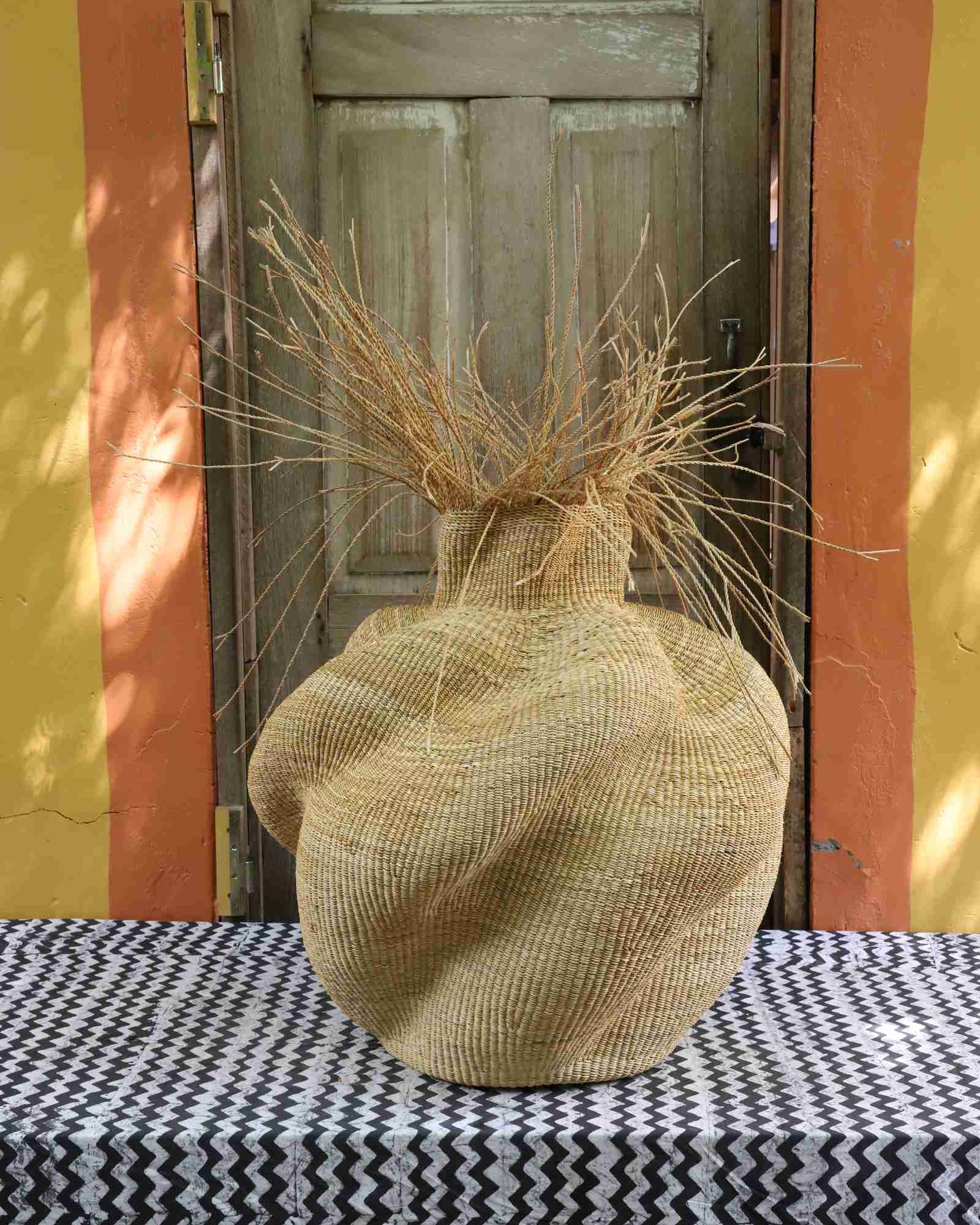 Bulbous Bolga basket in natural straw, its narrow neck topped with loose straw fronds, styled on a patterned tablecloth.