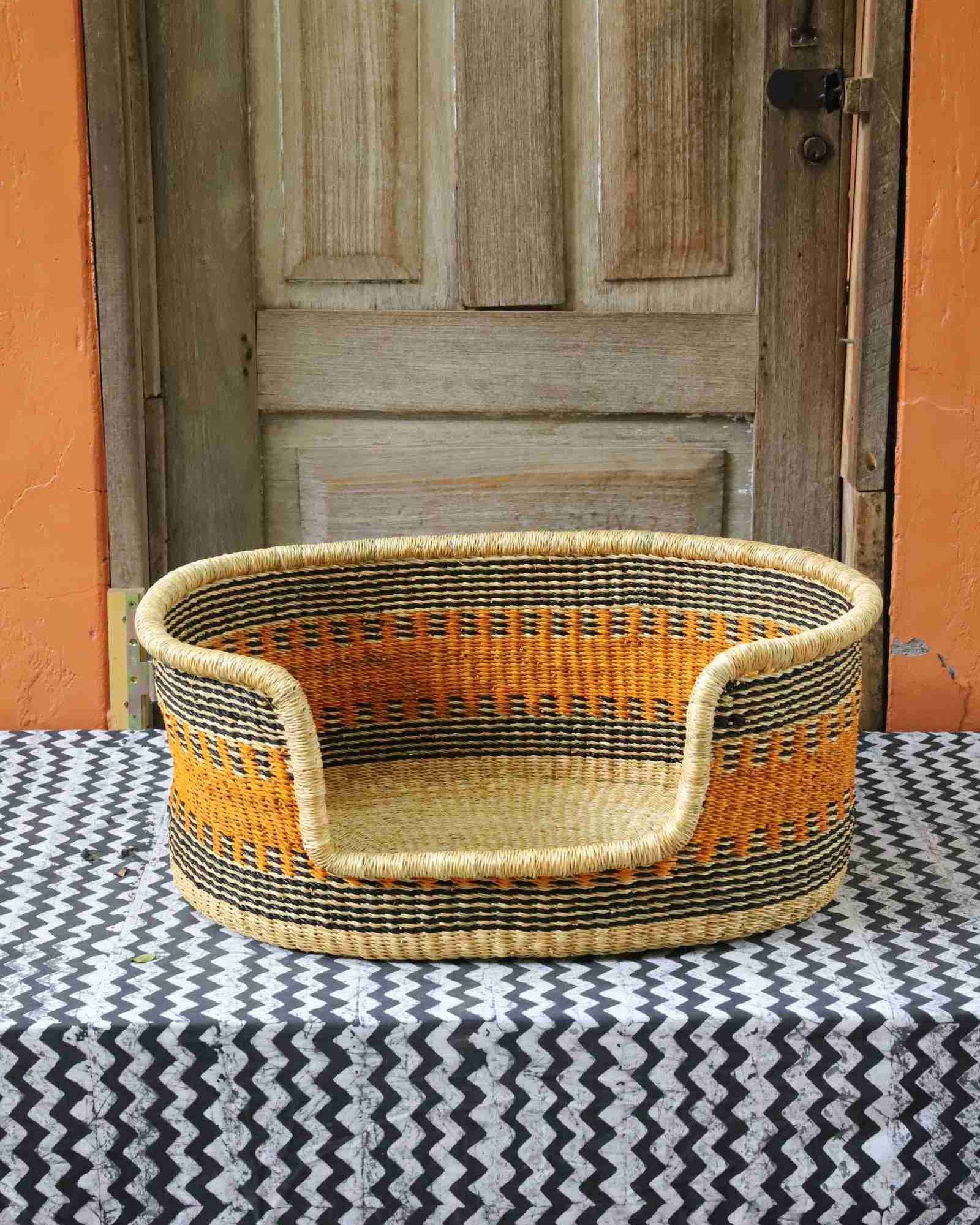 Woven dog bed in natural straw with an orange and dark checkered pattern, styled on a chevron cloth by a wooden door.