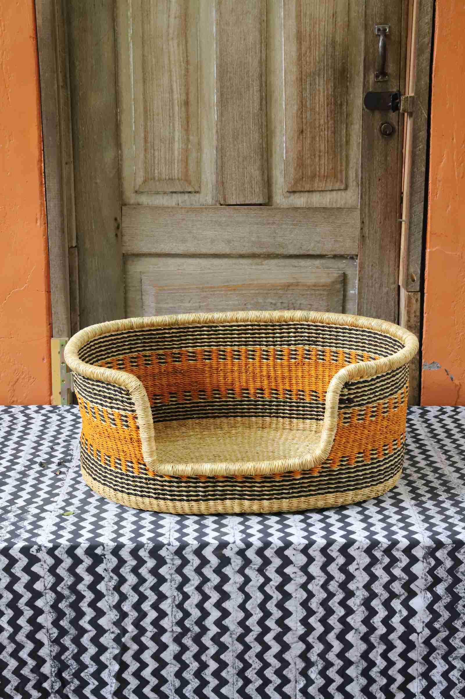 Woven dog bed in natural straw with an orange and dark checkered pattern, styled on a chevron cloth by a wooden door.
