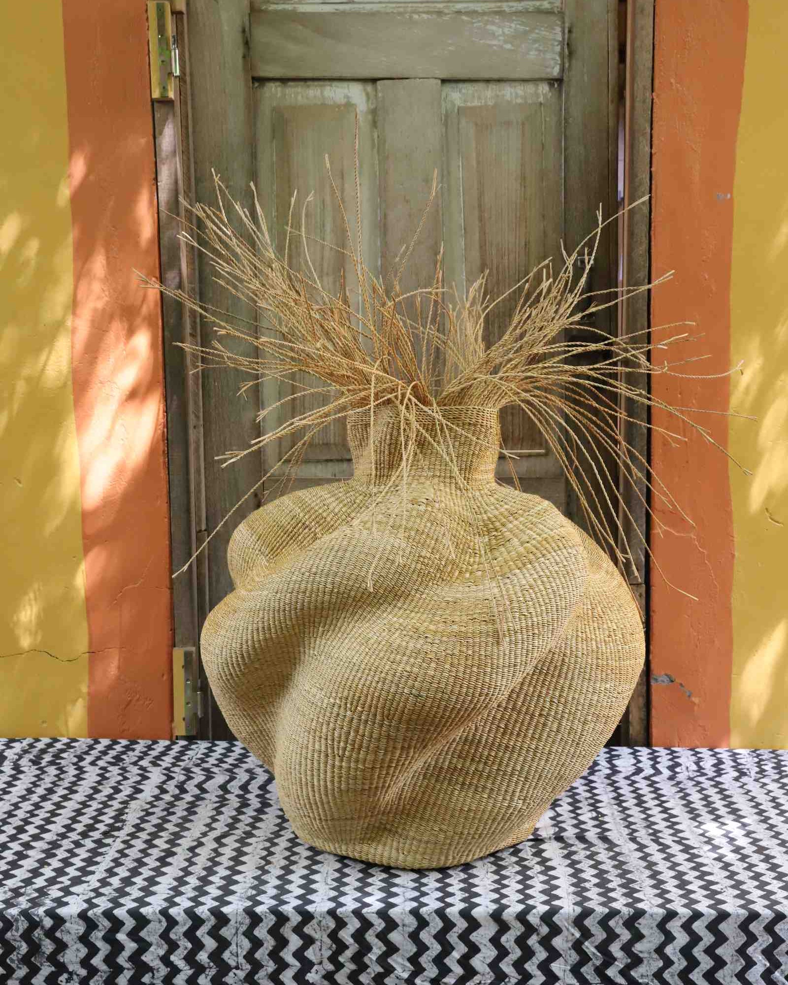Round Bolga basket in natural straw with a bulbous woven body and loose straw fronds at the neck.