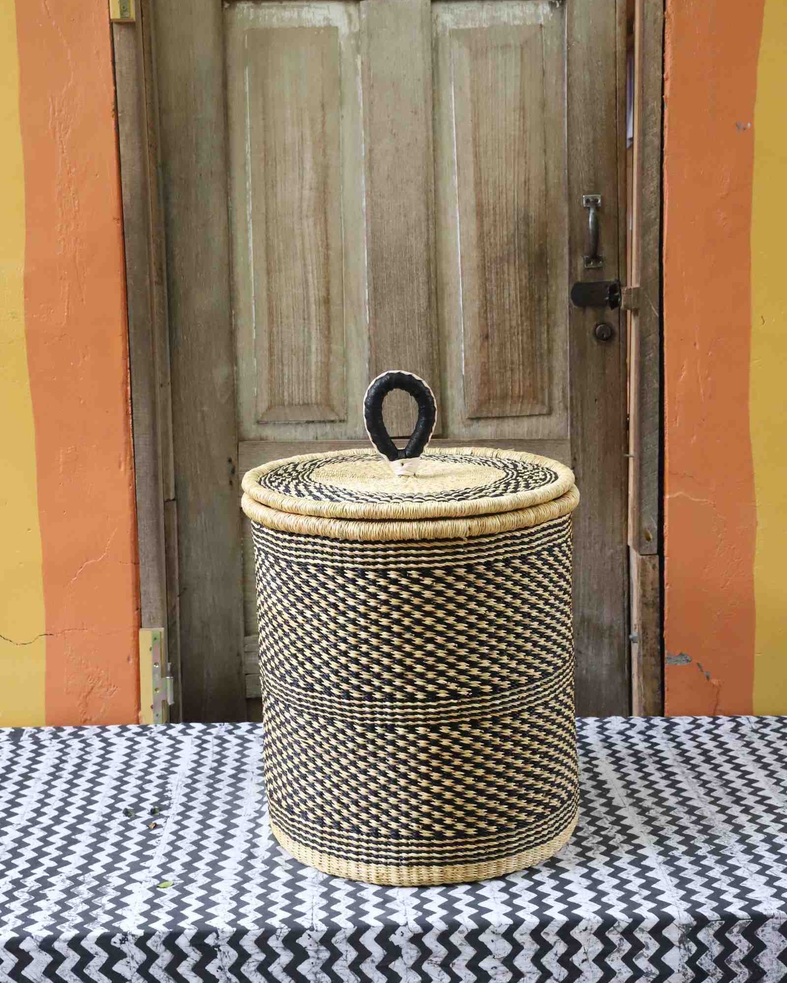 Coiled-rim lidded laundry basket in a black and tan speckled weave, styled on a chevron cloth by a wooden door.