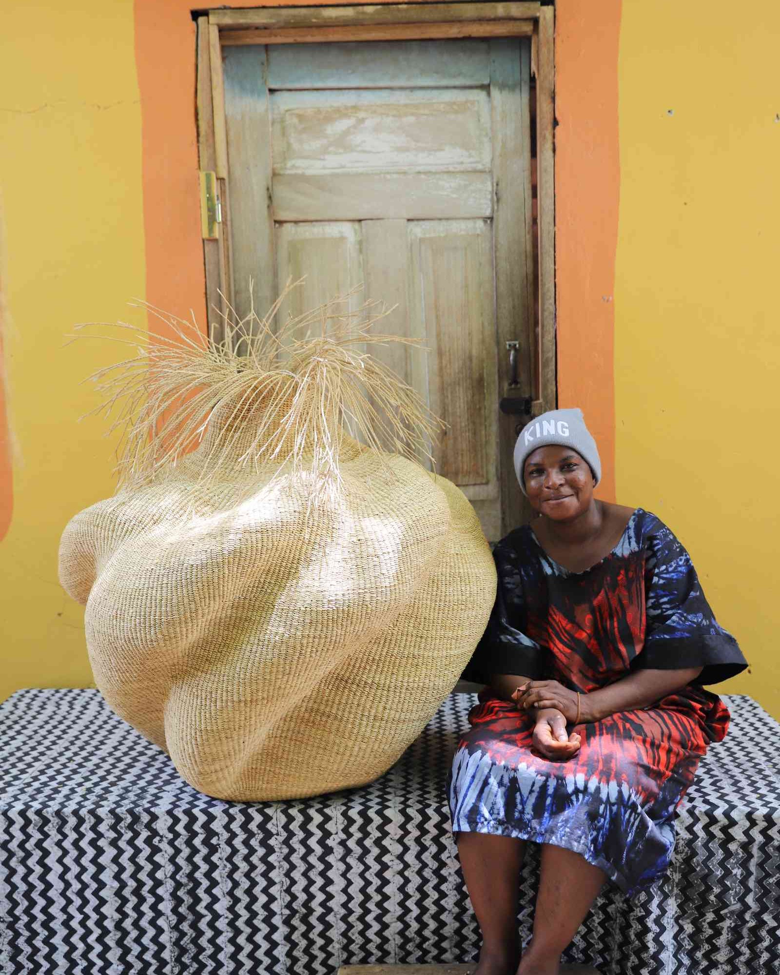 Handwoven art basket in natural straw with organic lobed body and dried grass stems, beside a woman seated in Bolgatanga.