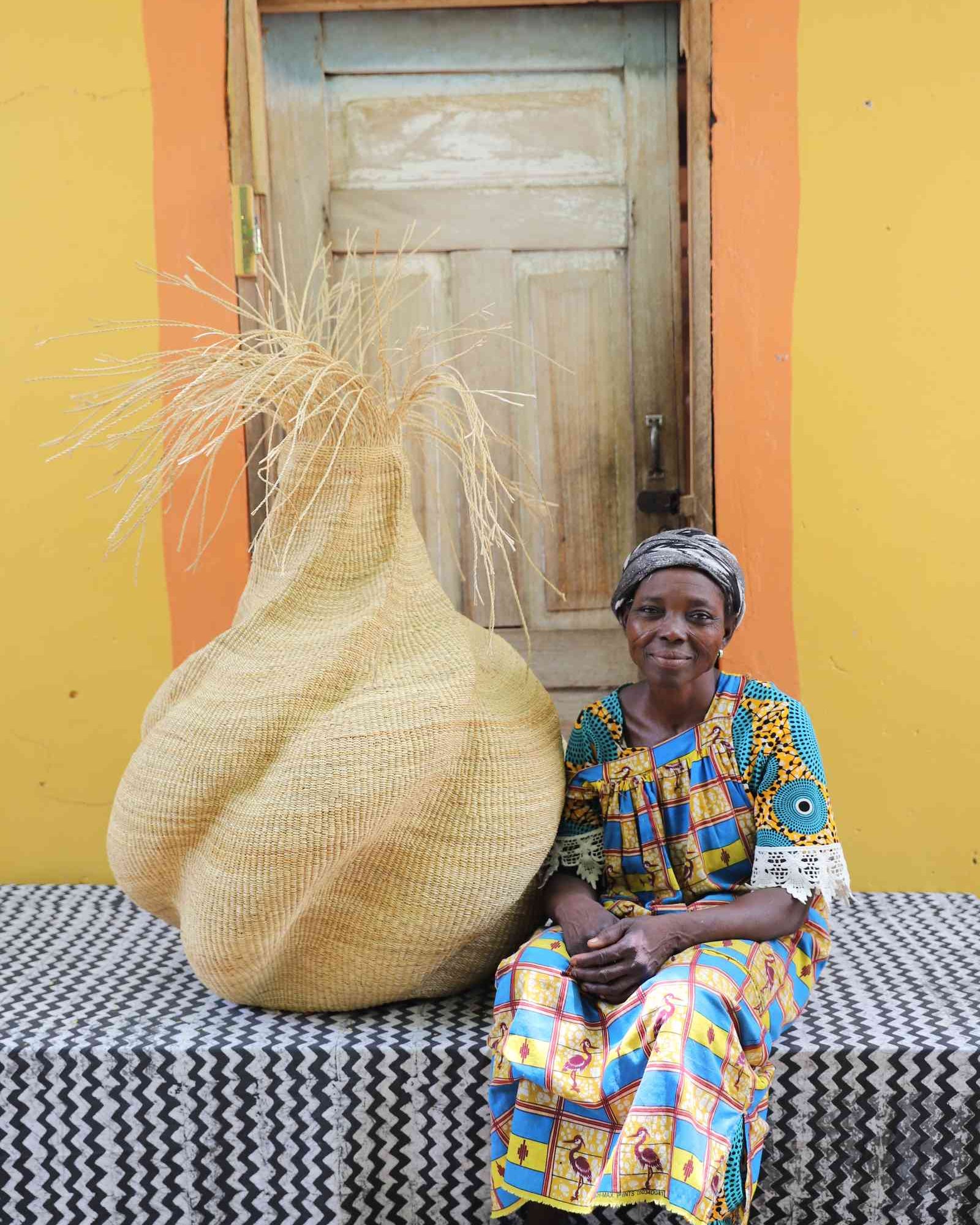 Handwoven art basket in natural straw with sculptural twisted body and dried grass stems, beside a woman in Bolgatanga.