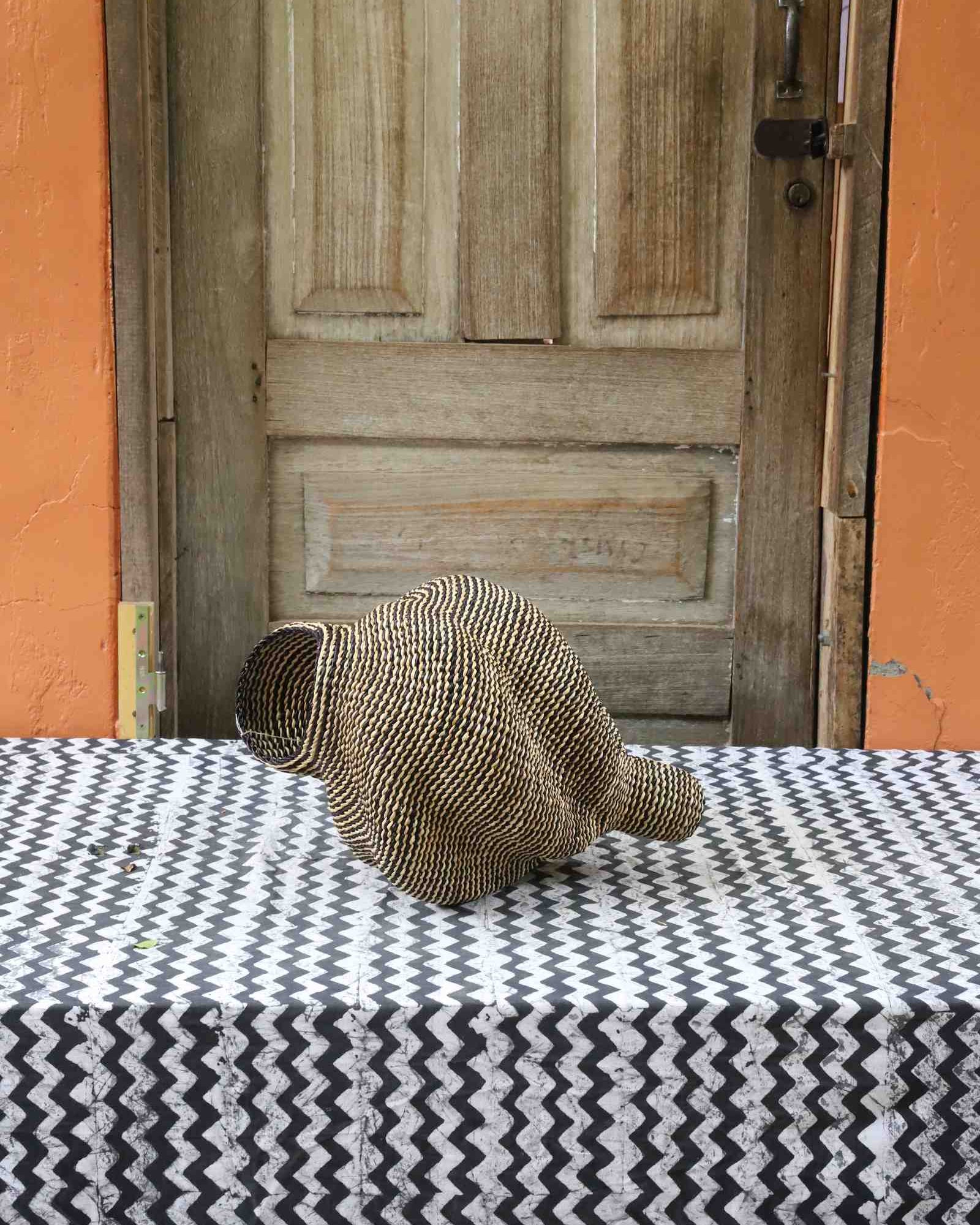 Small spiral-form Bolga art basket in natural straw, side view on a chevron cloth by a wooden door.