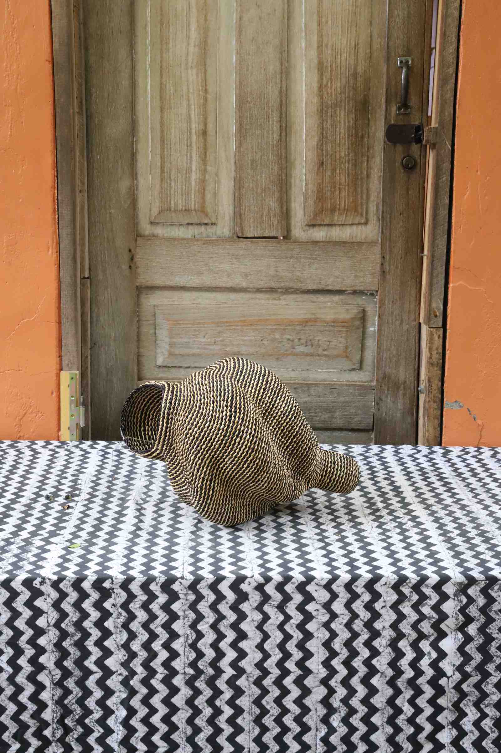 Small spiral-form Bolga art basket in natural straw, side view on a chevron cloth by a wooden door.