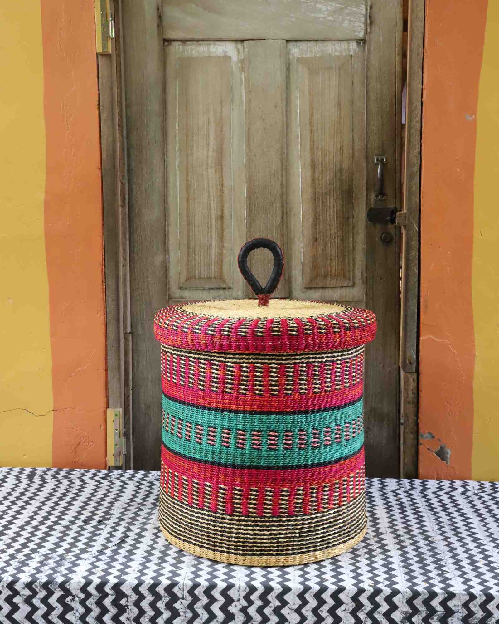 Handwoven Bolga laundry basket with lid in natural straw with bold red and green horizontal bands.