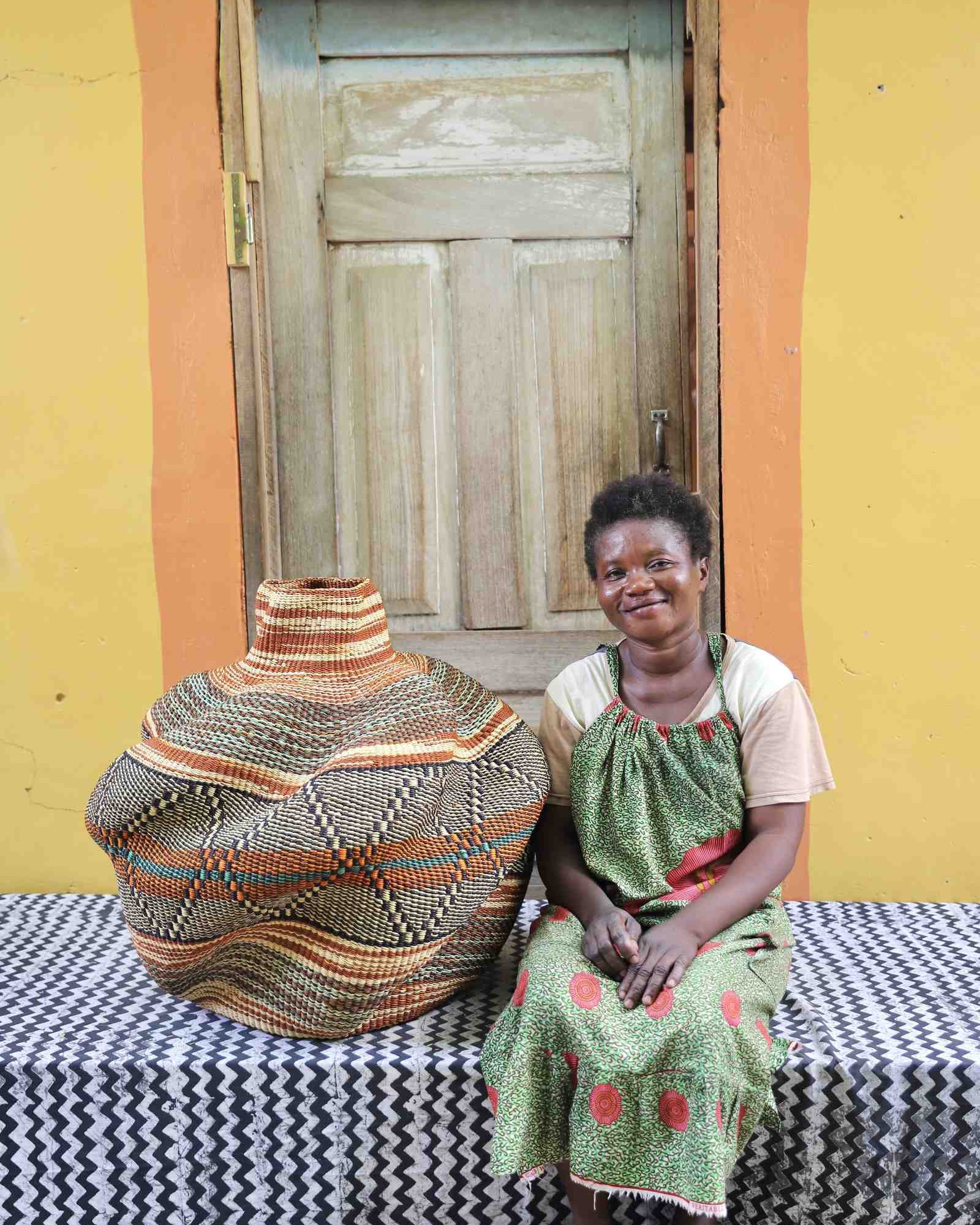 Handwoven African art basket in sculptural lobed vase form with earth-tone stripes, beside a smiling woman in Bolgatanga.