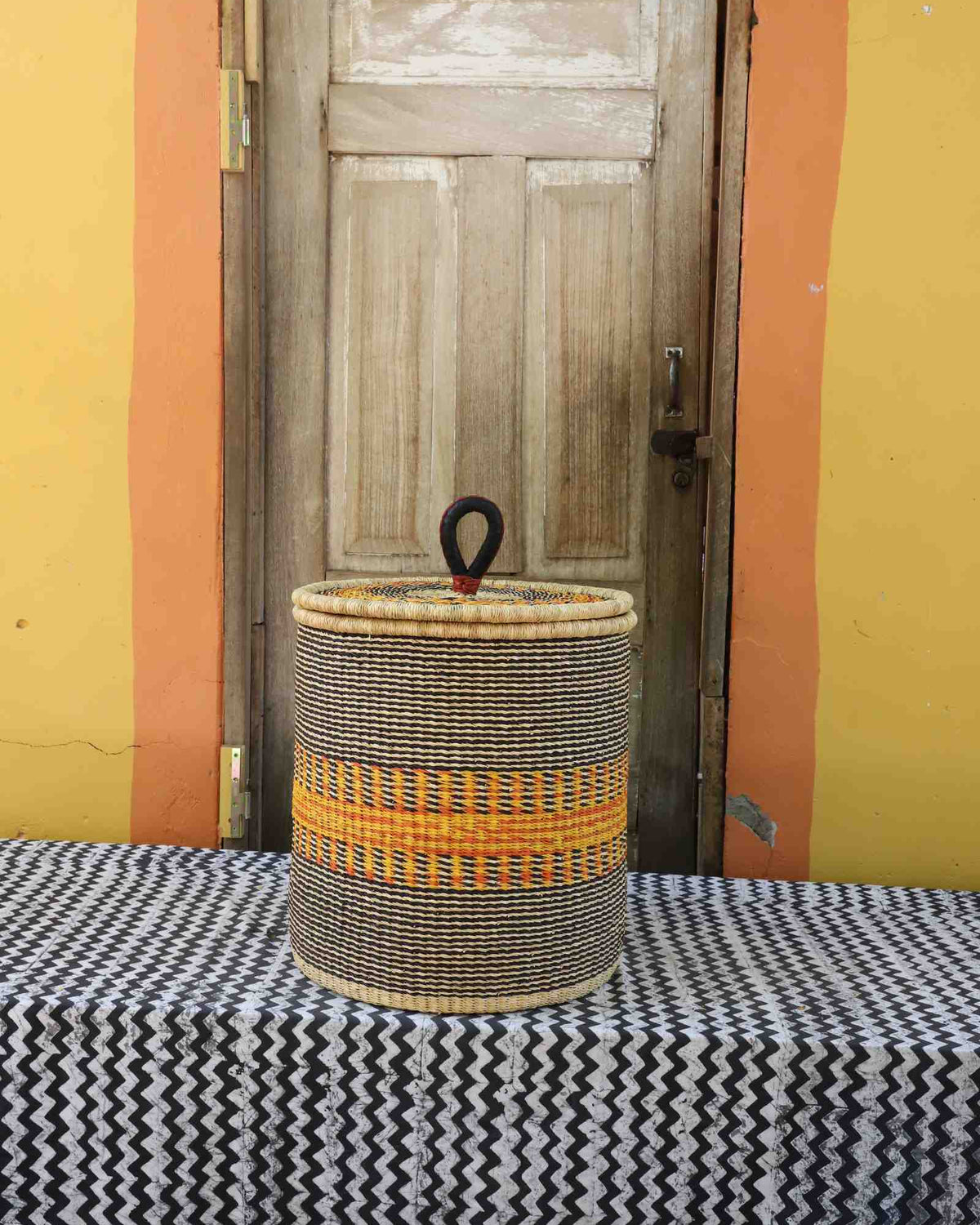 Handwoven Bolga laundry basket with coiled rim and lid in natural straw with a bold orange central band.