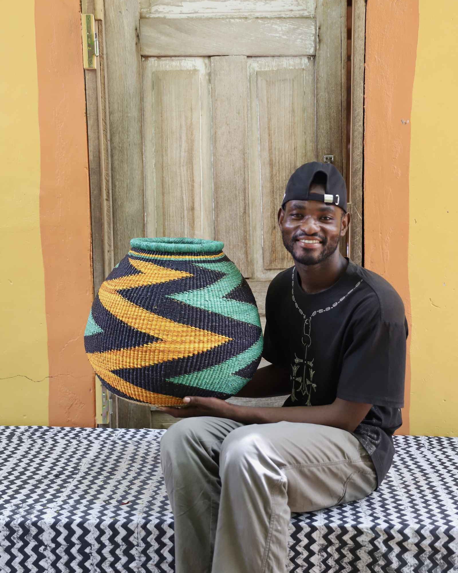 Handwoven African art basket with bold diagonal zigzag pattern in teal, black and amber, held by a man in Bolgatanga.