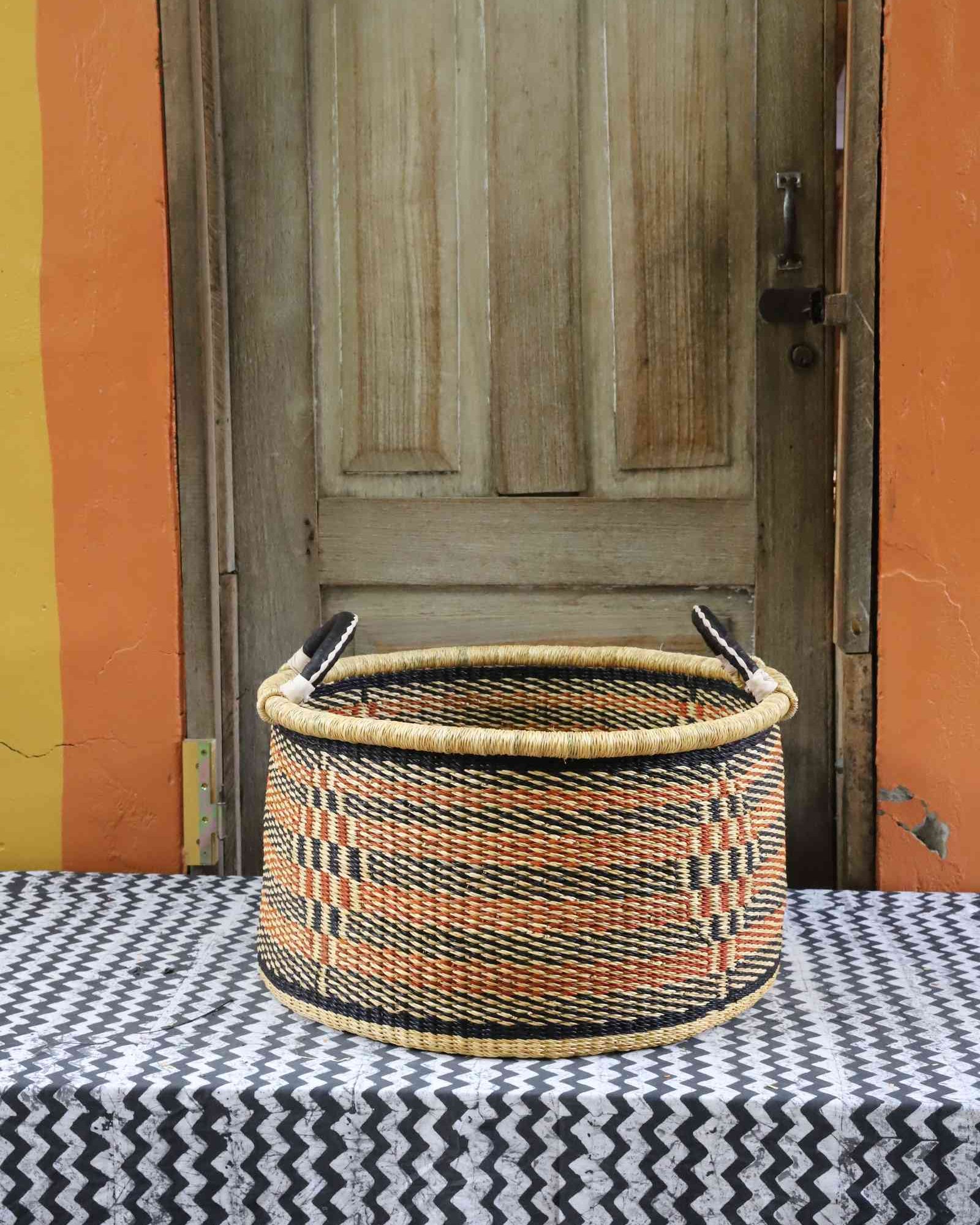 Handwoven Bolga storage basket in natural straw with orange and black horizontal stripes and twin handles.