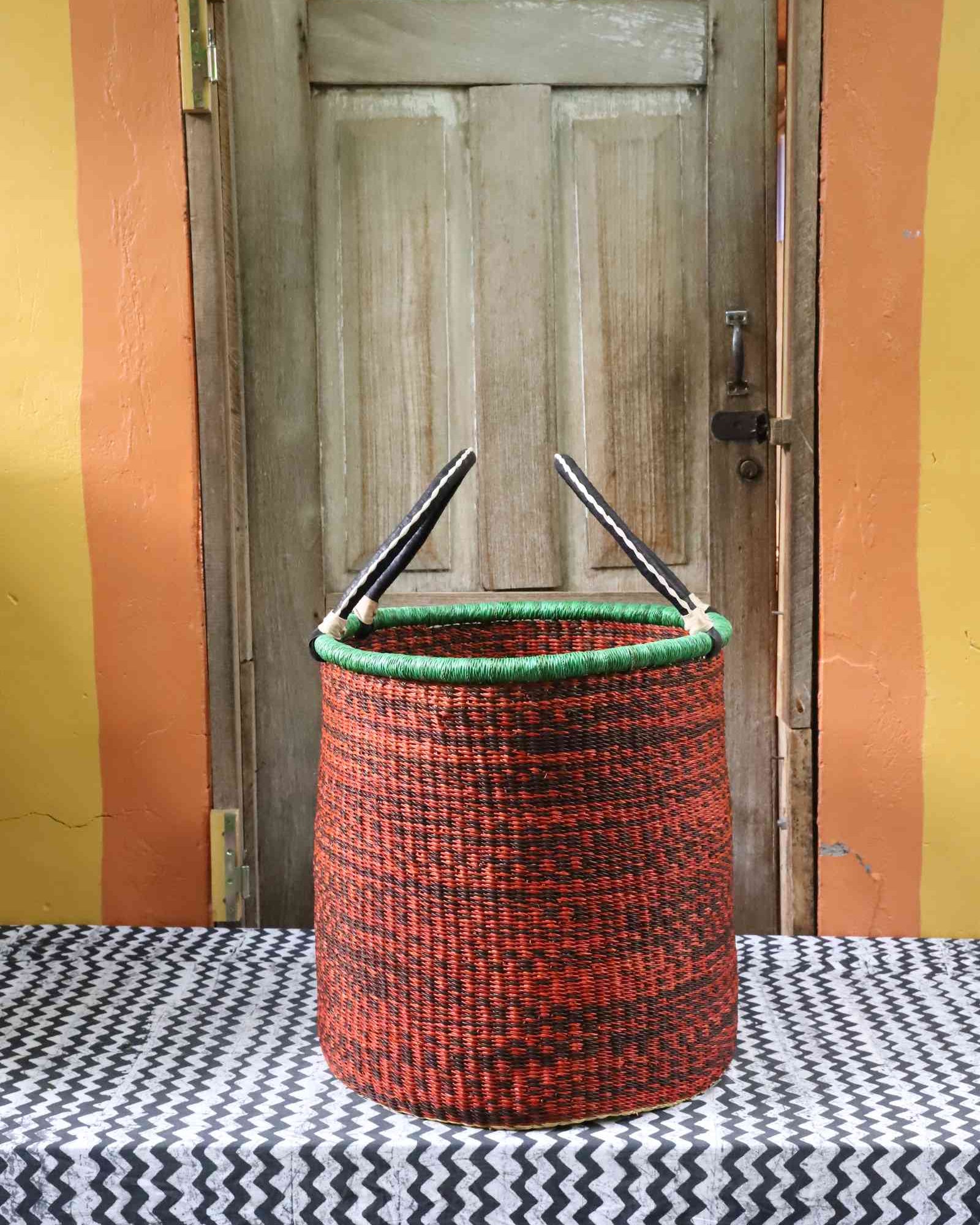 Tall open Bolga laundry basket in speckled rust-red weave with a green rim edge and leather-topped handles.