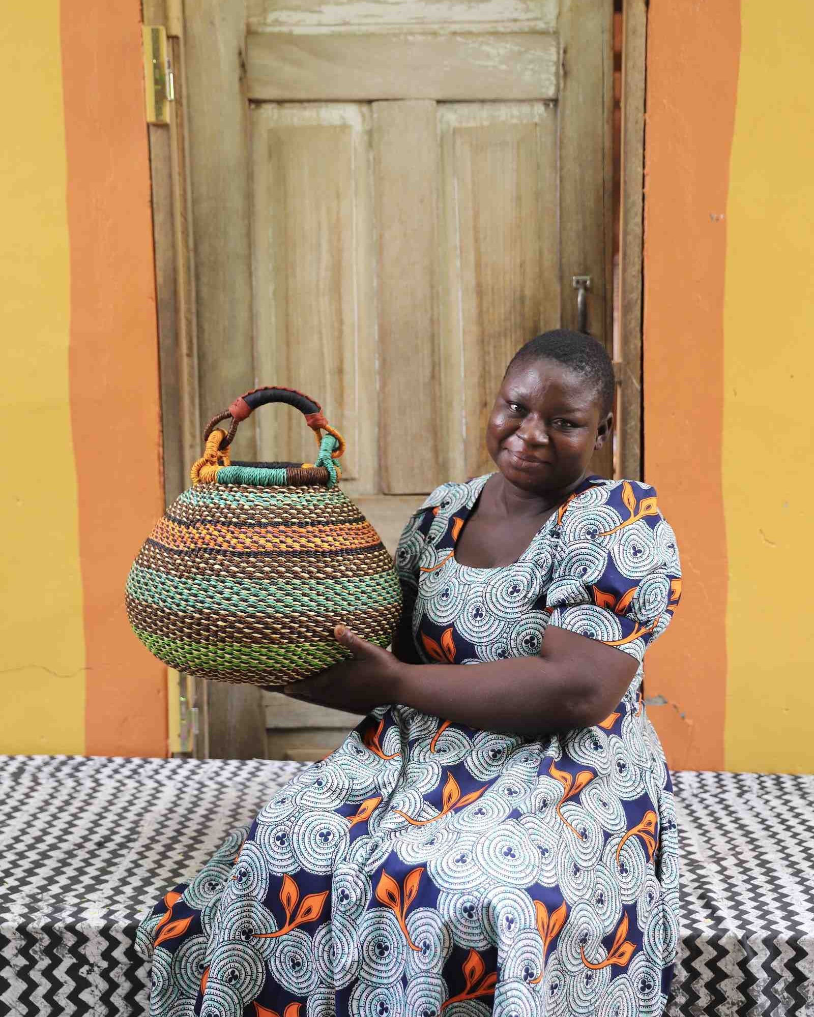Handwoven Bolga pot basket in multicoloured bands of green, teal and amber, held by a woman seated in Bolgatanga.