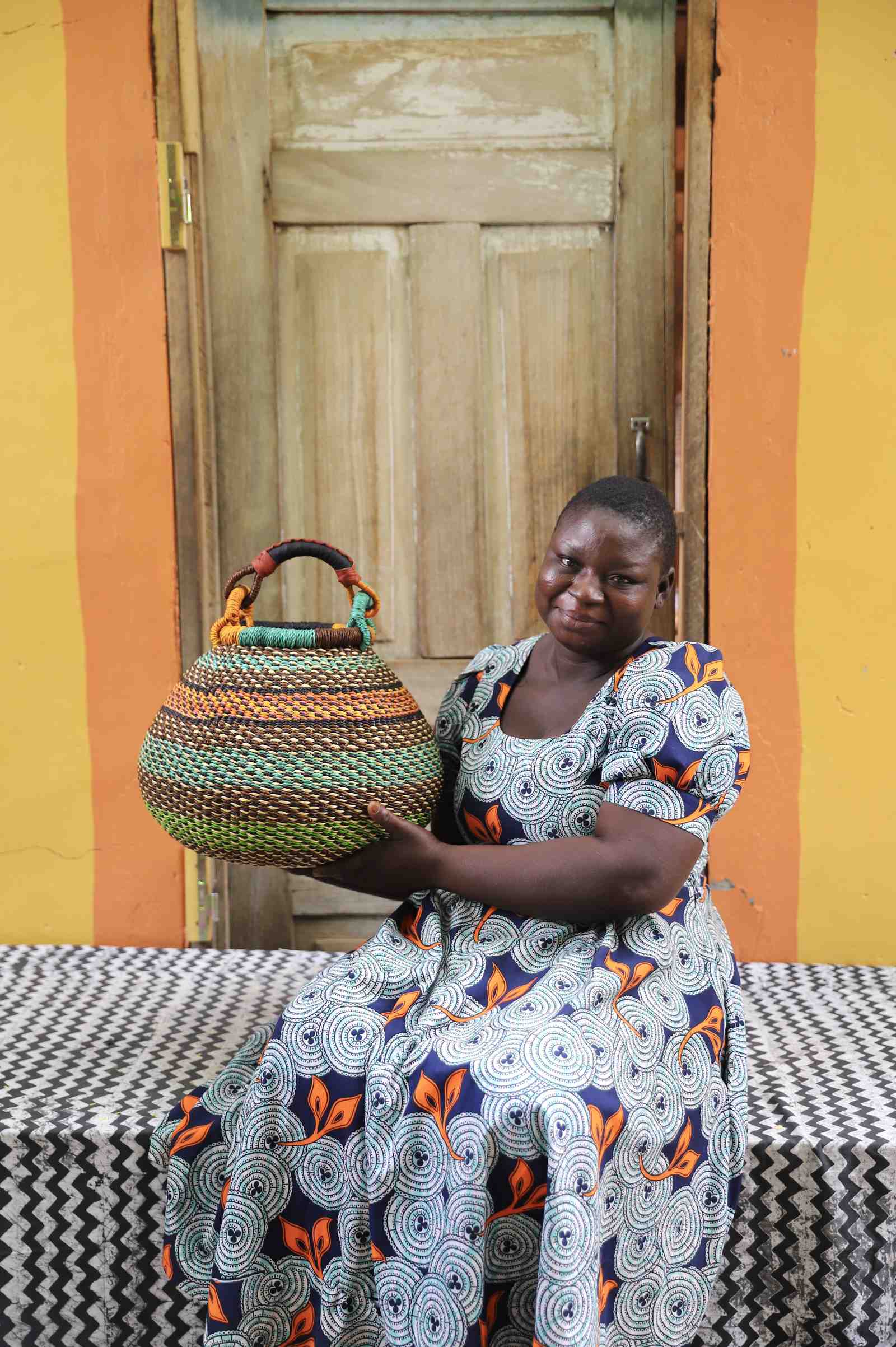 Handwoven Bolga pot basket in multicoloured bands of green, teal and amber, held by a woman seated in Bolgatanga.