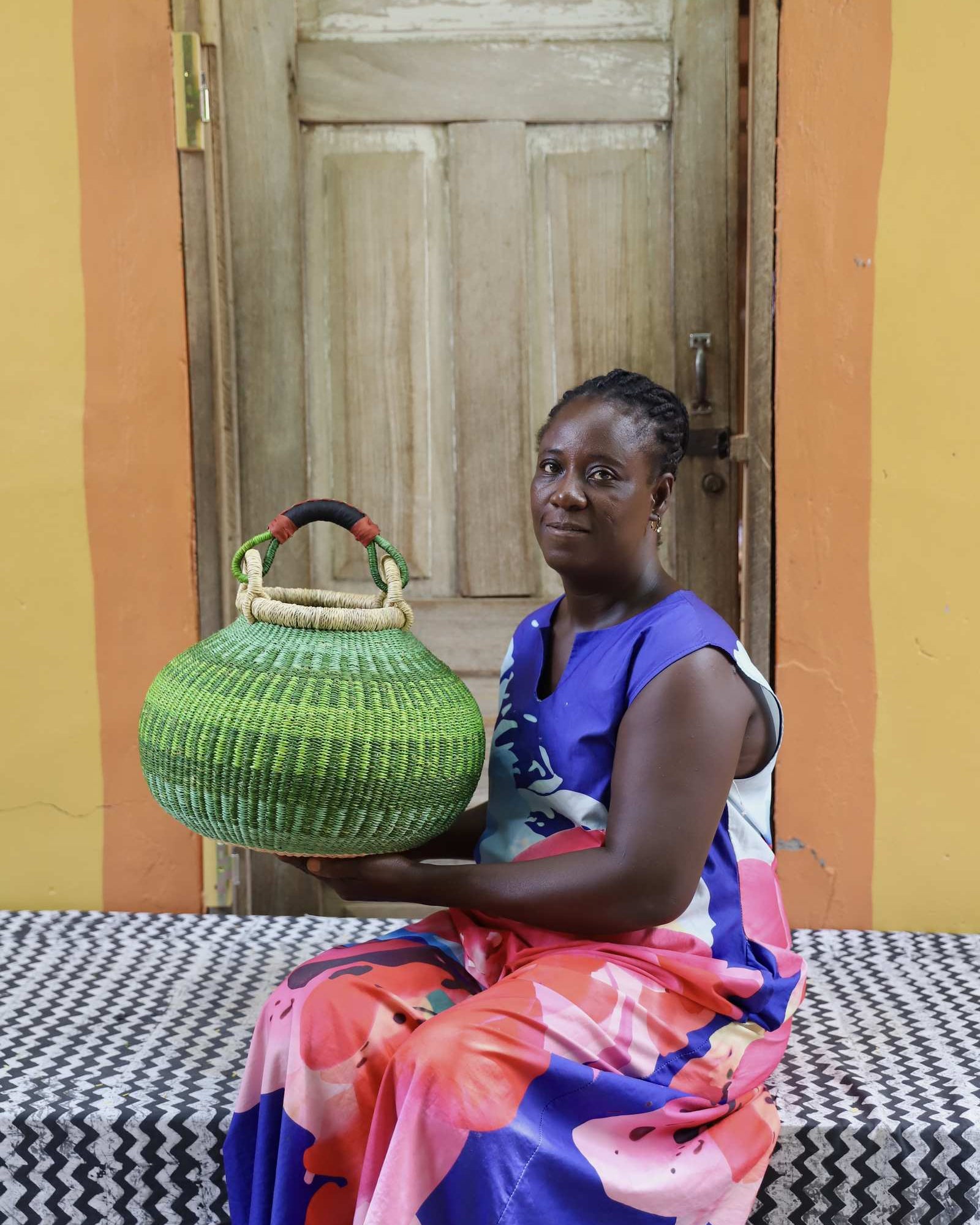 Large handwoven Bolga pot basket in lime green elephant grass with leather top handle, held by a woman seated in Bolgatanga.