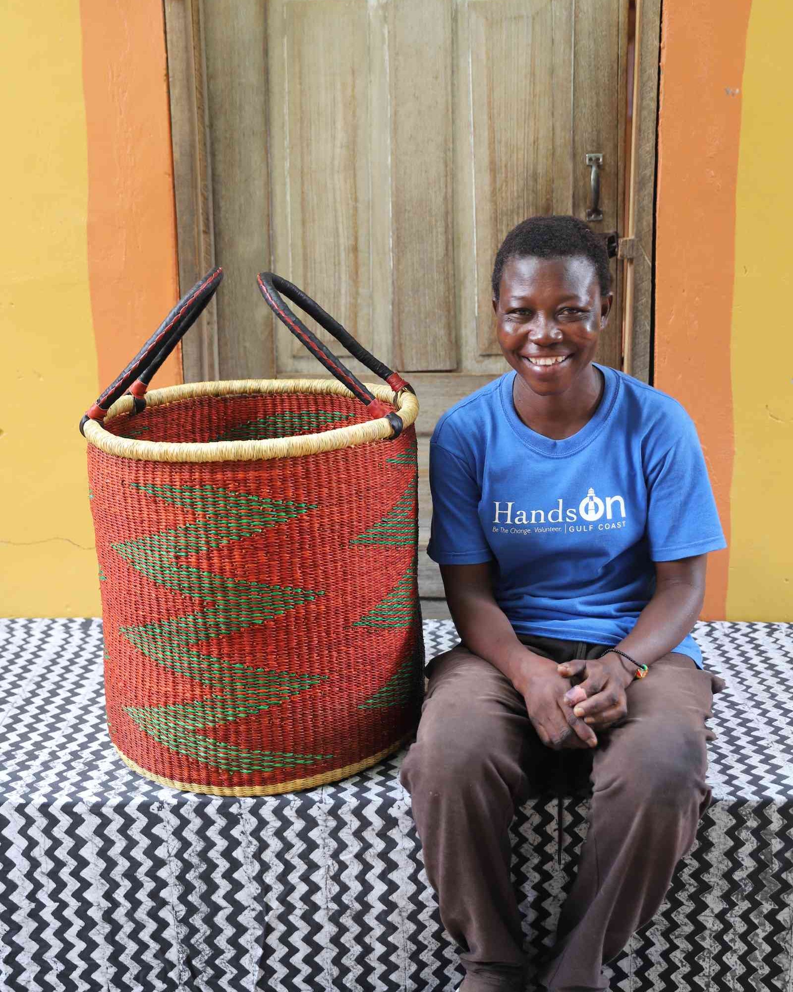 Large open laundry basket in red and green zigzag weave, shown beside a seated person for scale.