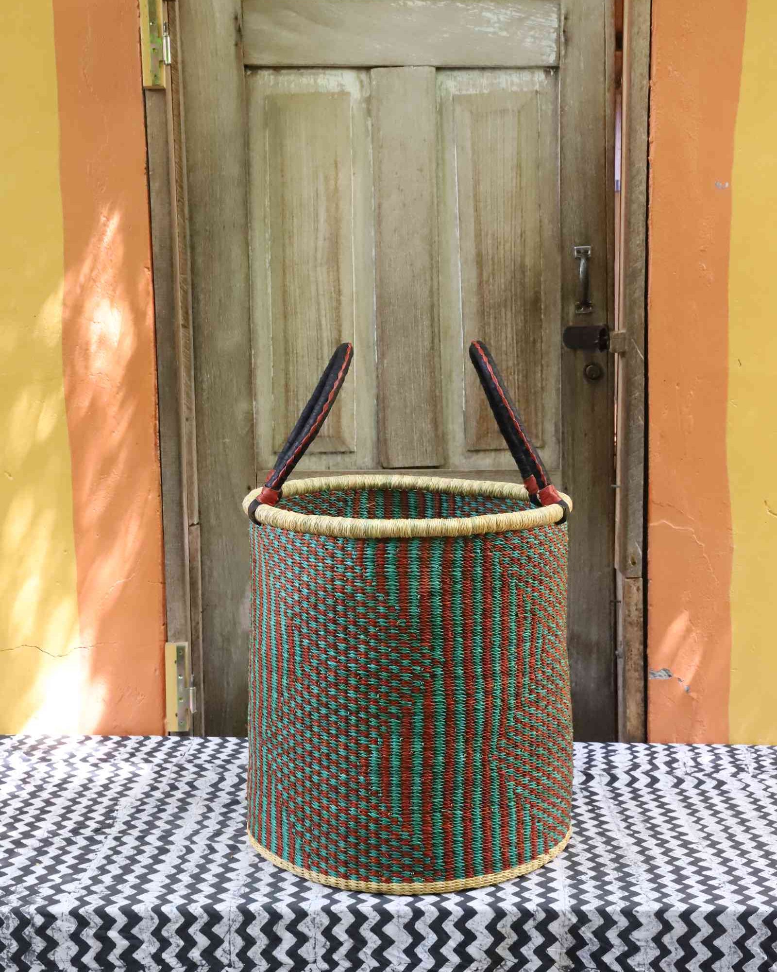 Tall open Bolga laundry basket in a green and rust striped weave with leather-topped handles, on a tablecloth.