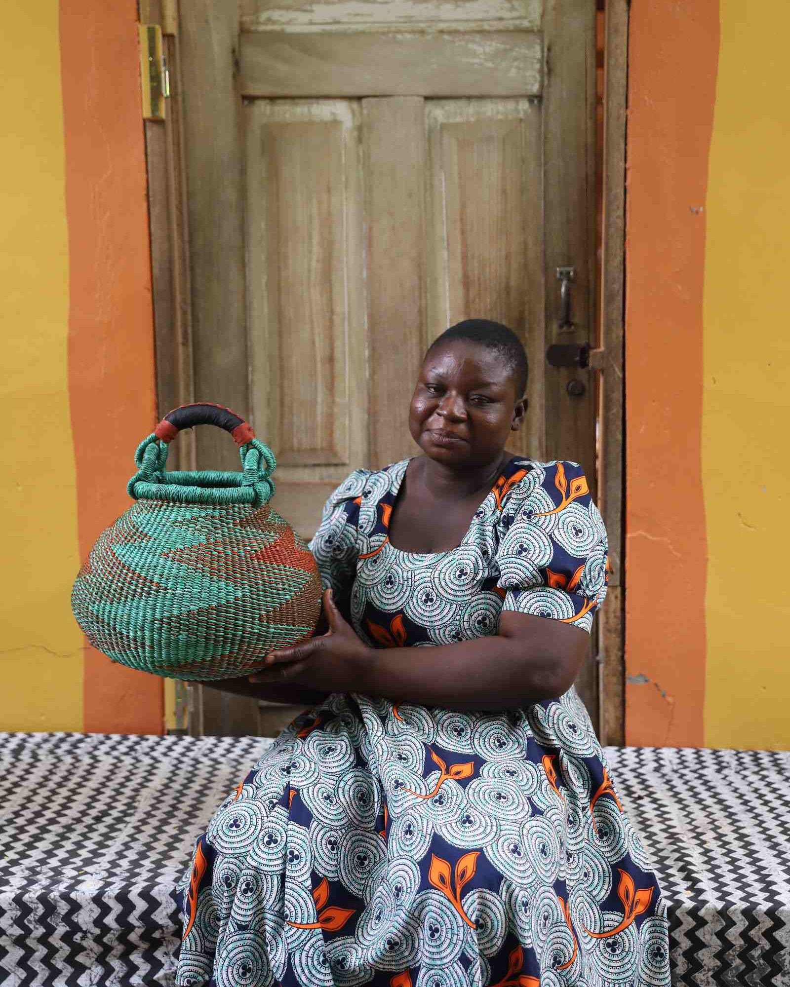 Small handwoven Bolga pot basket in teal and terracotta with leather top handle, held by a woman in Bolgatanga.