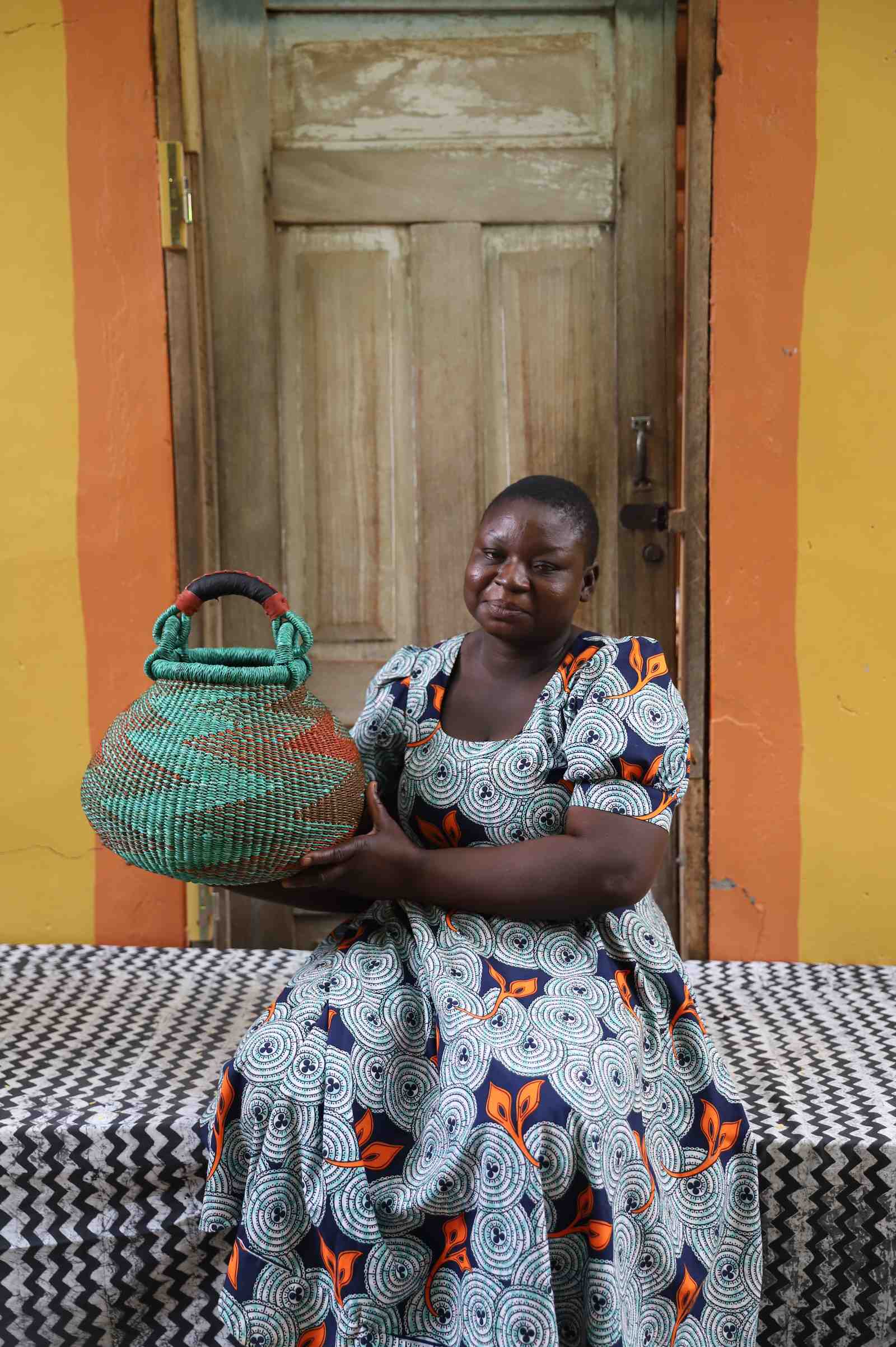 Small handwoven Bolga pot basket in teal and terracotta with leather top handle, held by a woman in Bolgatanga.