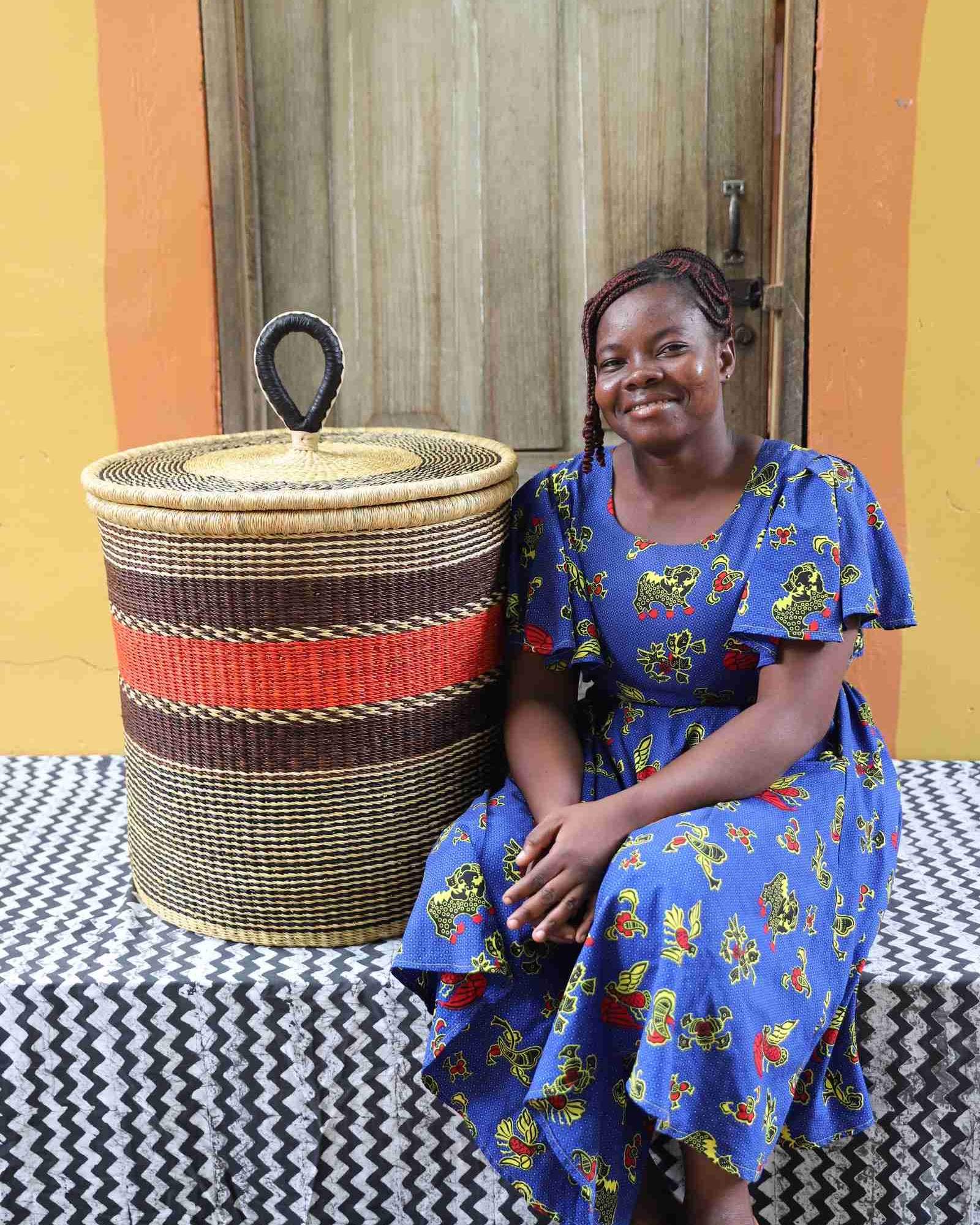 Large coiled-rim lidded laundry basket in natural straw with a red and brown band, shown beside a seated person for scale.